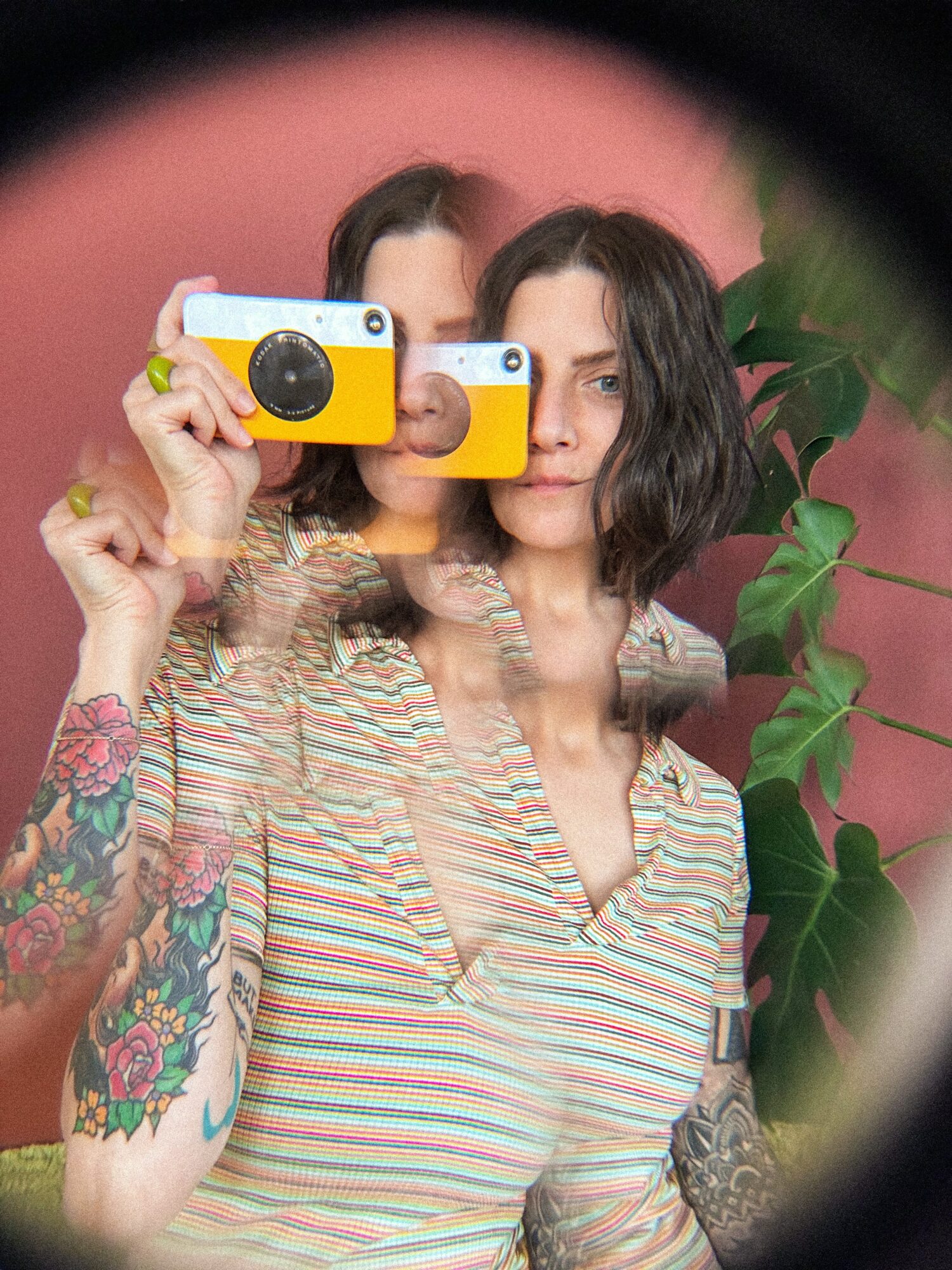 Two women with dark hair take a selfie with yellow cameras, standing in front of a pink background with green leaves.