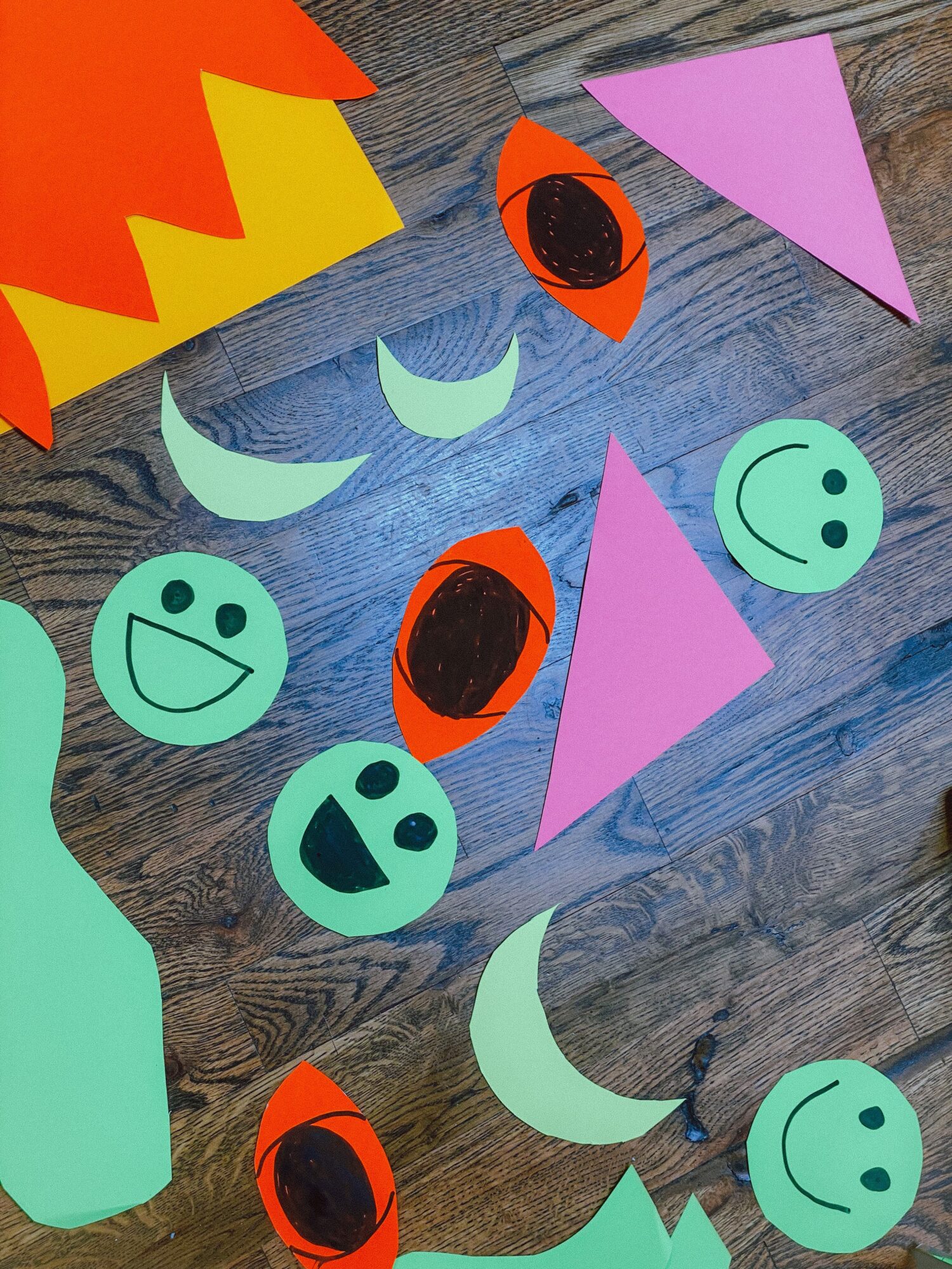 Colorful paper cutouts of faces, shapes, and a moon on a wooden surface, arranged randomly.