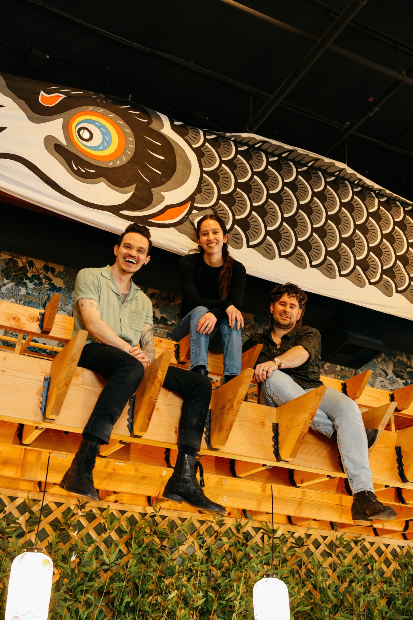 Three people sitting on wooden benches in front of a large dragon mural with scales and a fierce face, smiling and posing.