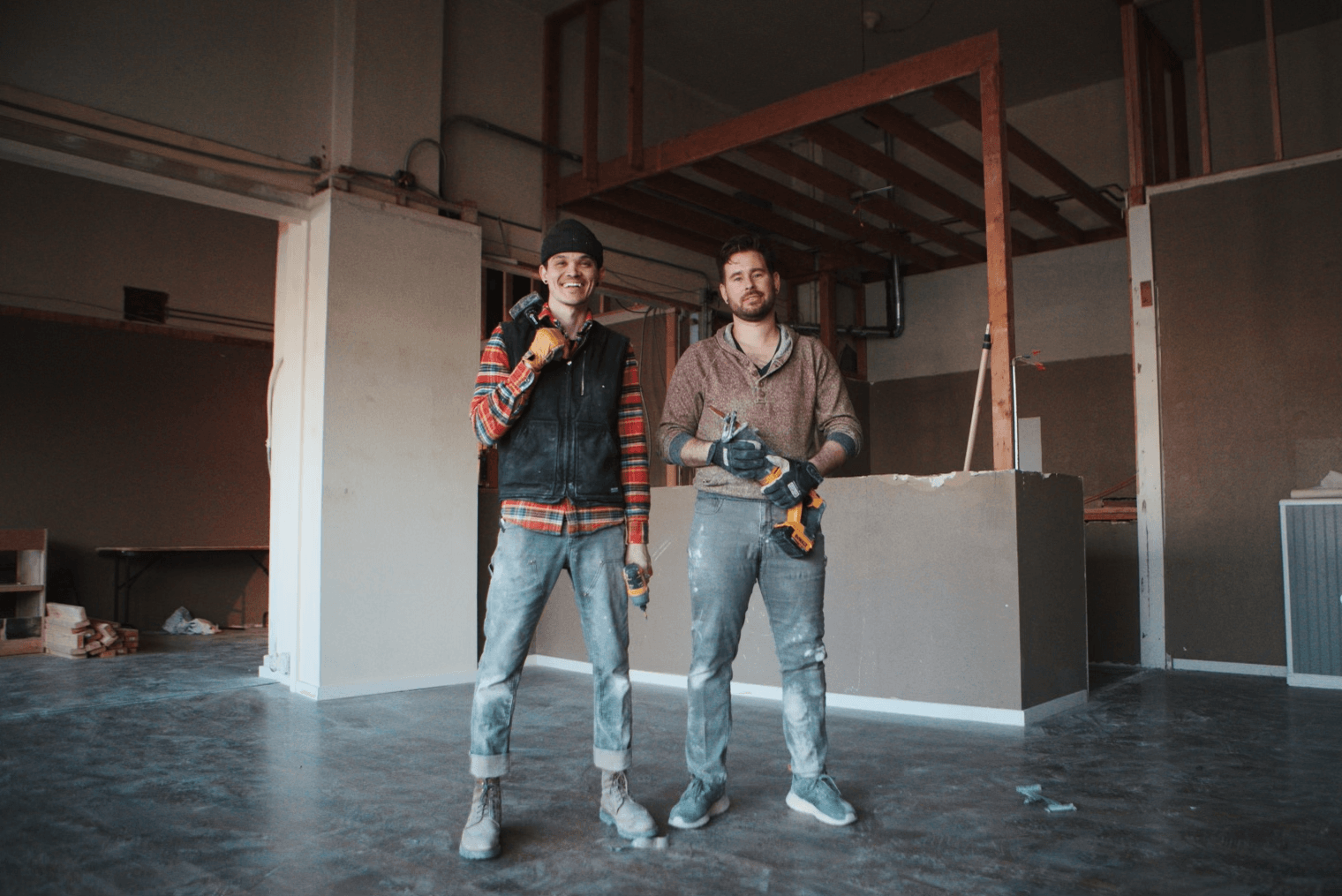 Two men stand inside a construction site, one holding a power drill, both dressed casually. Construction materials are visible around them.