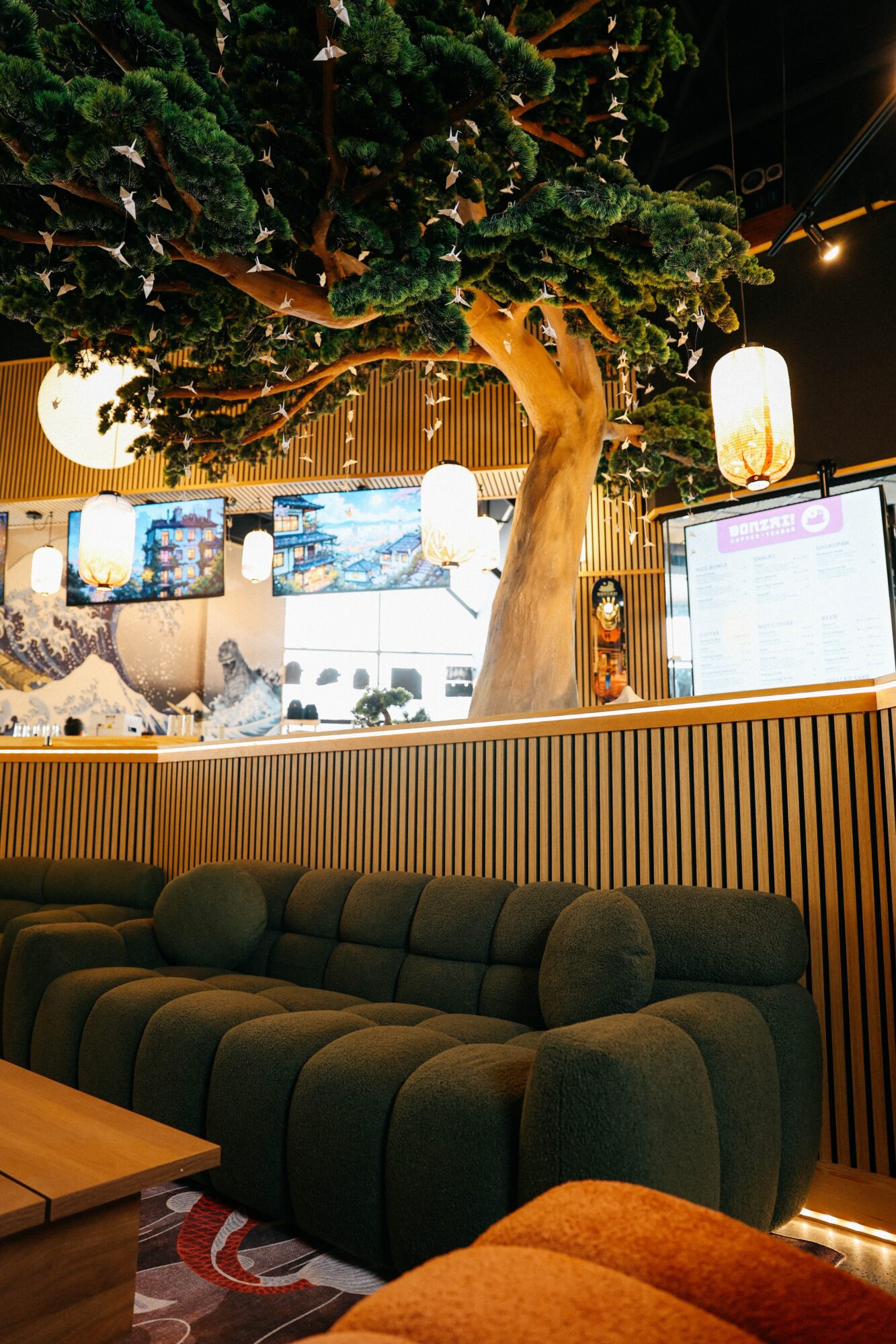 Indoor space with a large artificial tree, illuminated lamps, a digital screen, and a dark green sofa with cushions.