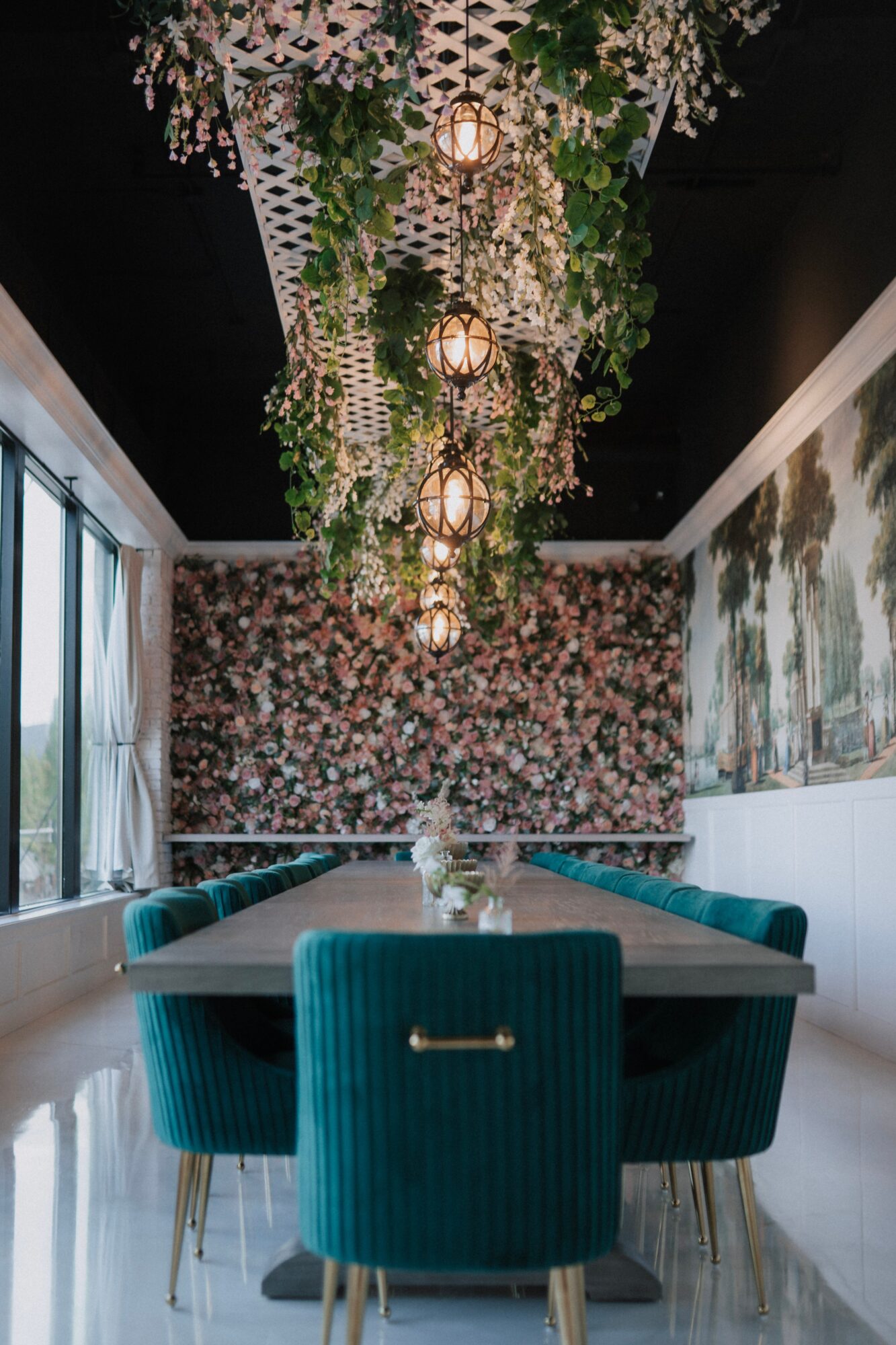 Round table with teal chairs in a room with floral wallpaper and a floral ceiling installation with hanging lights.