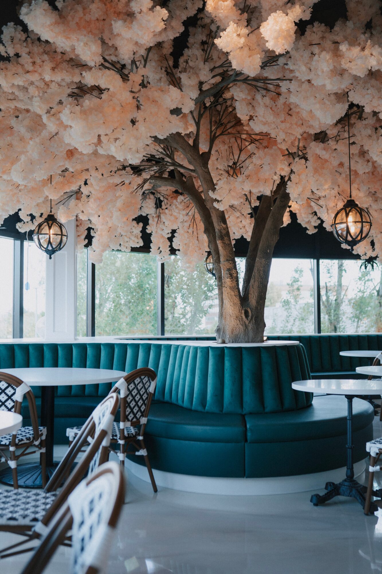 Indoor space with a large tree and pink blossoms, teal seating, and hanging lanterns, with windows showing outdoor trees.
