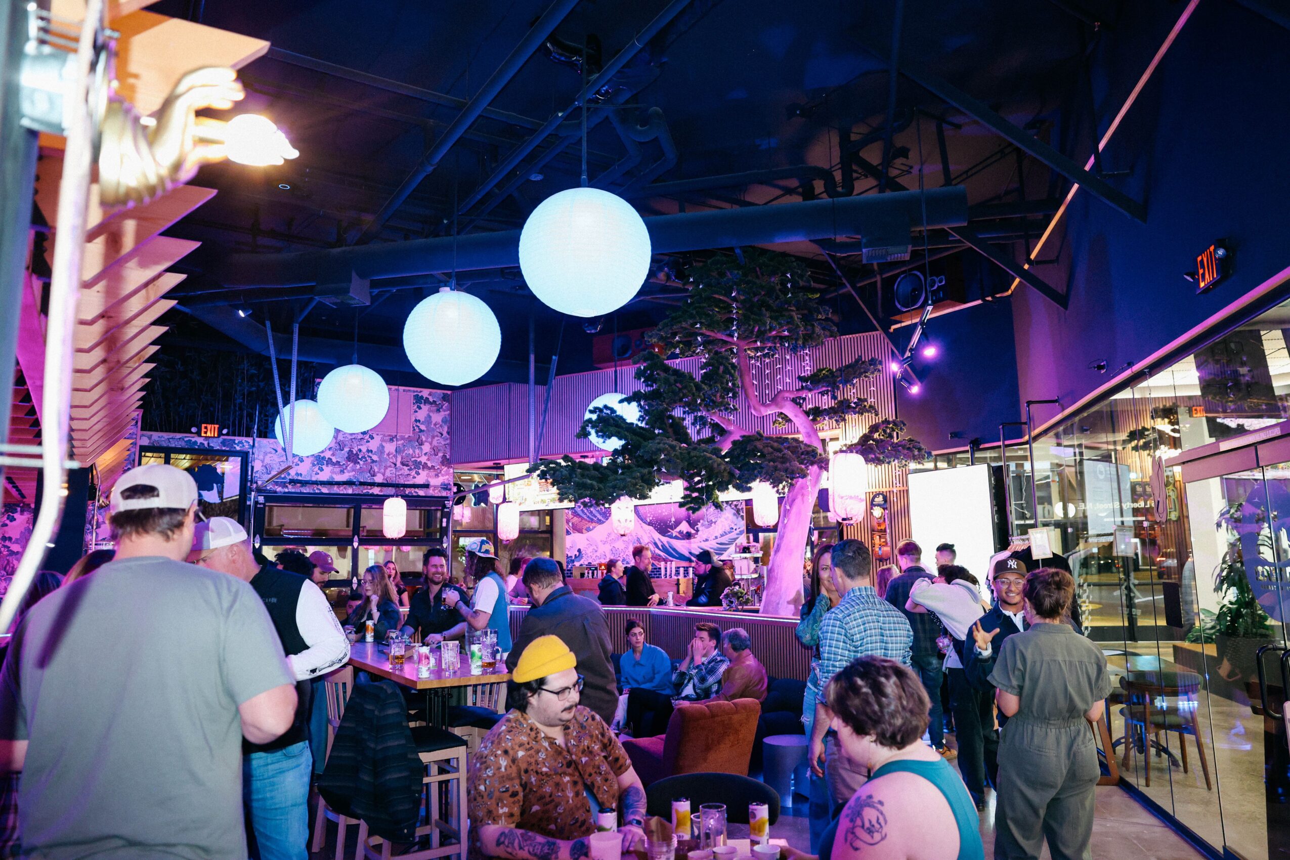 People gather in a lively indoor space with hanging spherical lights and a large tree, some seated and others walking around.
