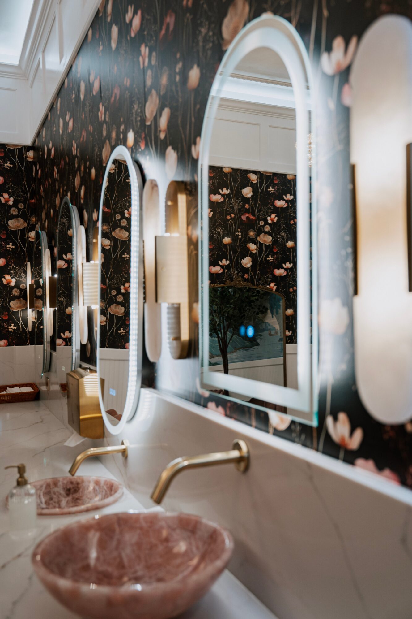 Bathroom with floral wallpaper, oval mirrors, pink marble sinks, and gold fixtures, illuminated by soft lighting.