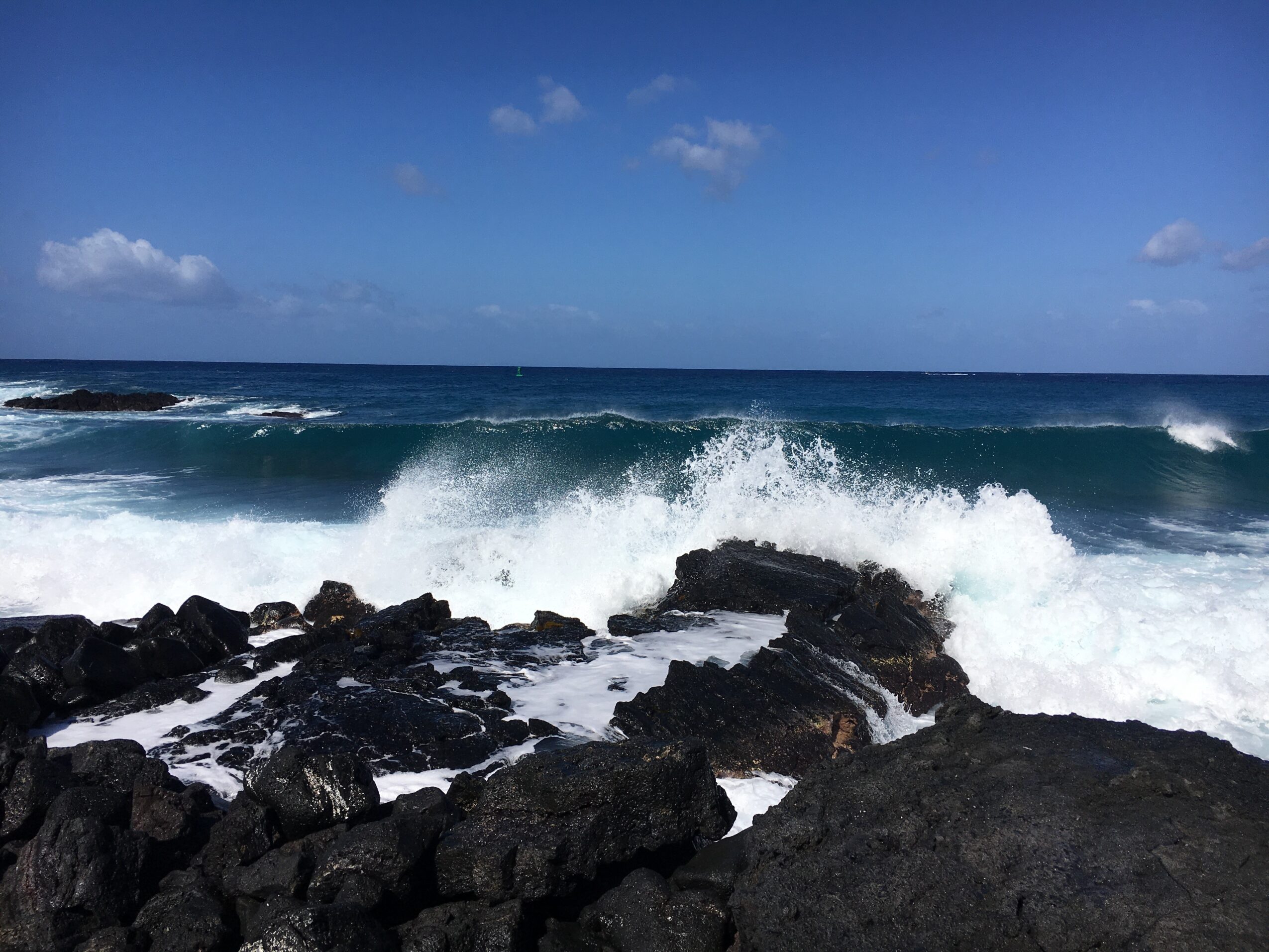 Ocean waves crashing against dark rocks under a blue sky with scattered clouds.