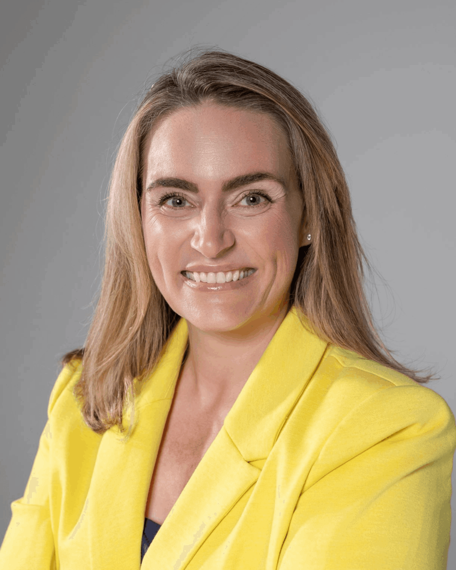 Woman with shoulder-length hair smiling, wearing a yellow blazer against a gray background.