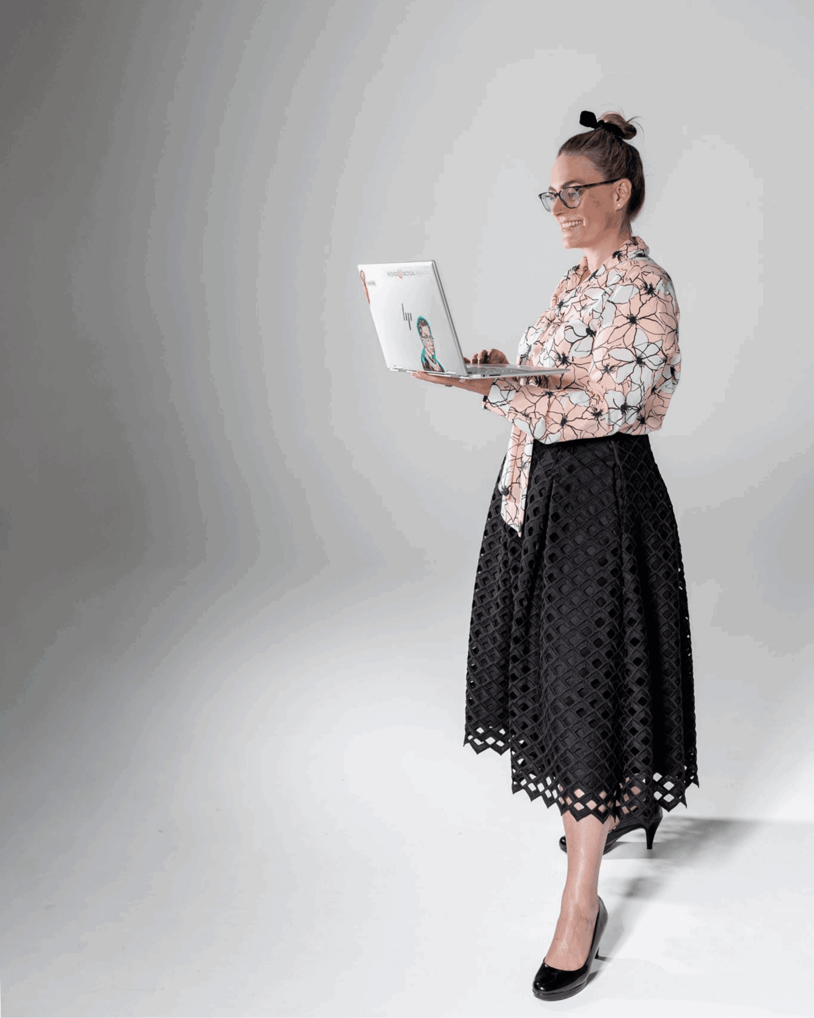 Woman with glasses and a bun hairstyle using a laptop, wearing a patterned blouse and a black lace skirt, standing against a plain background.