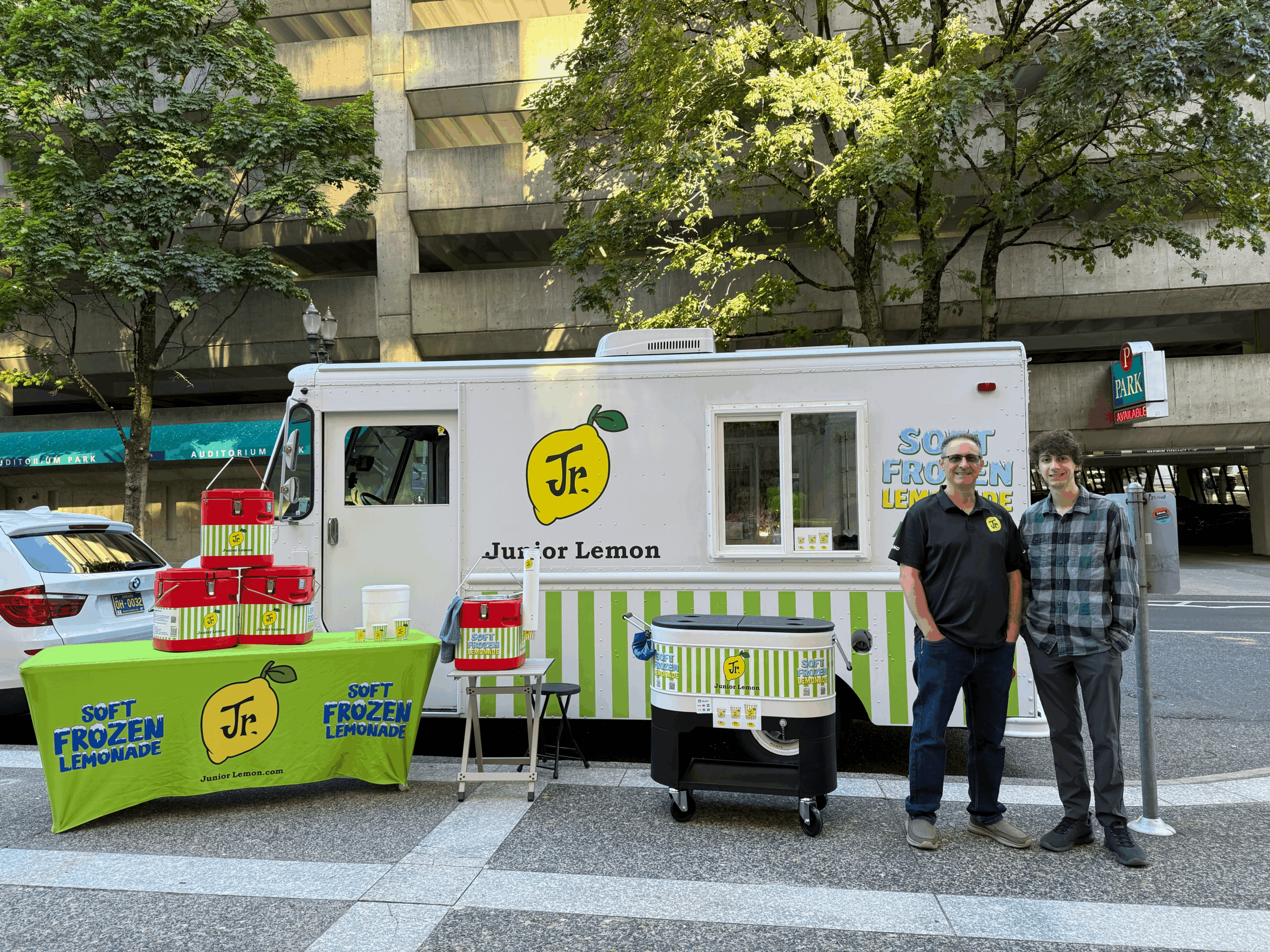 Two people stand in front of a mobile frozen lemonade stand with a white truck and green accents, on a city street.