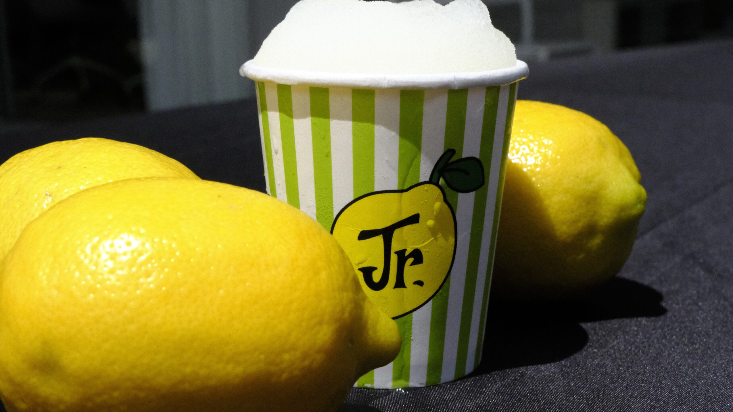 Two lemons and a cup with a lemon illustration and foam on top, on a dark surface.