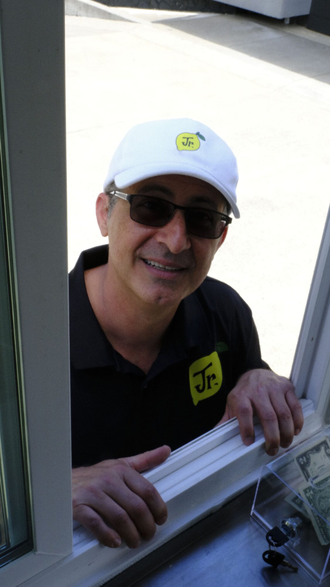 Person wearing sunglasses and a white cap, smiling, standing behind a counter with money and papers.