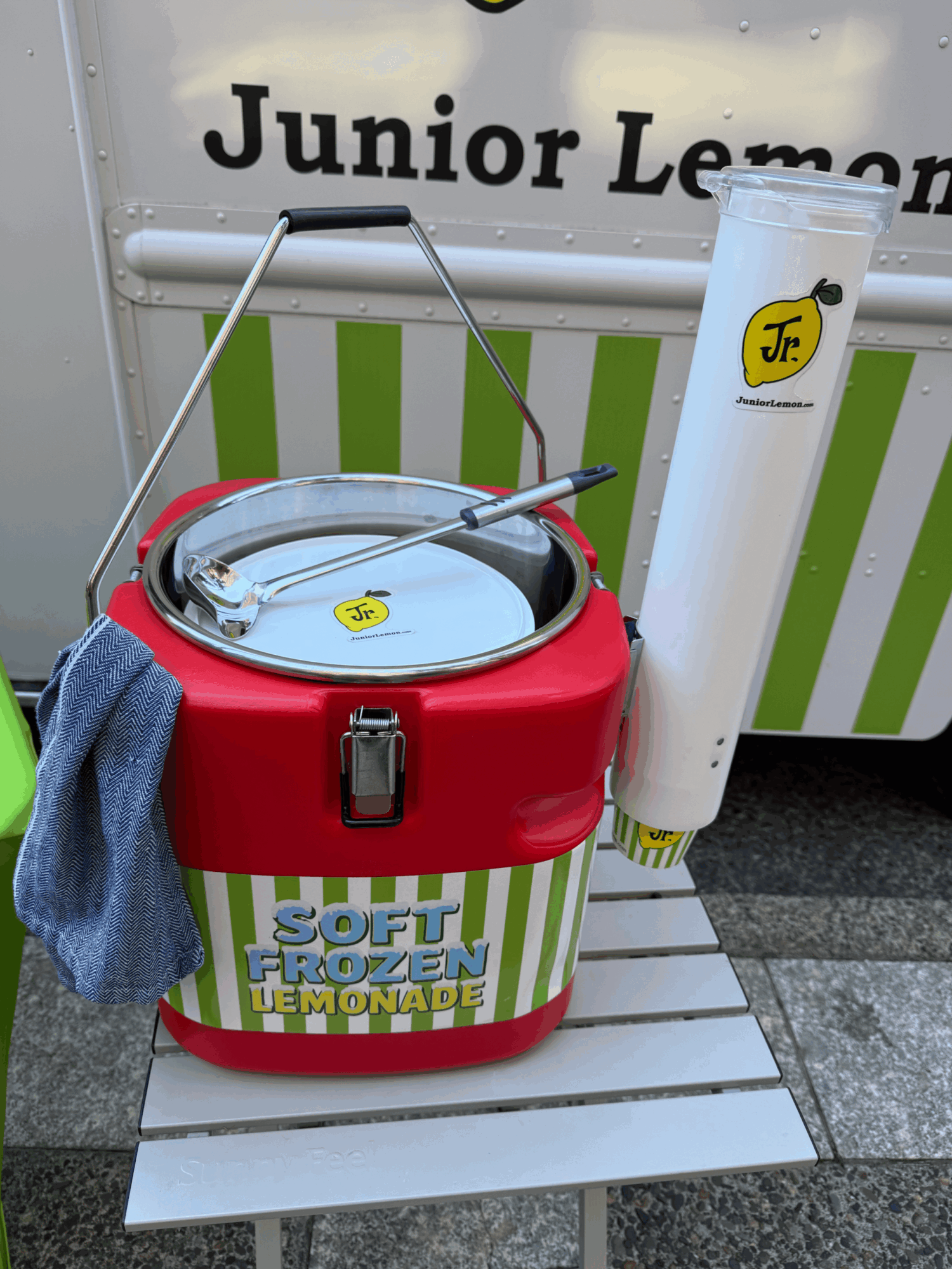 A red and white cooler with a towel hanging on it, placed on a white platform outside. A sign reads 'Junior Lemonade'.