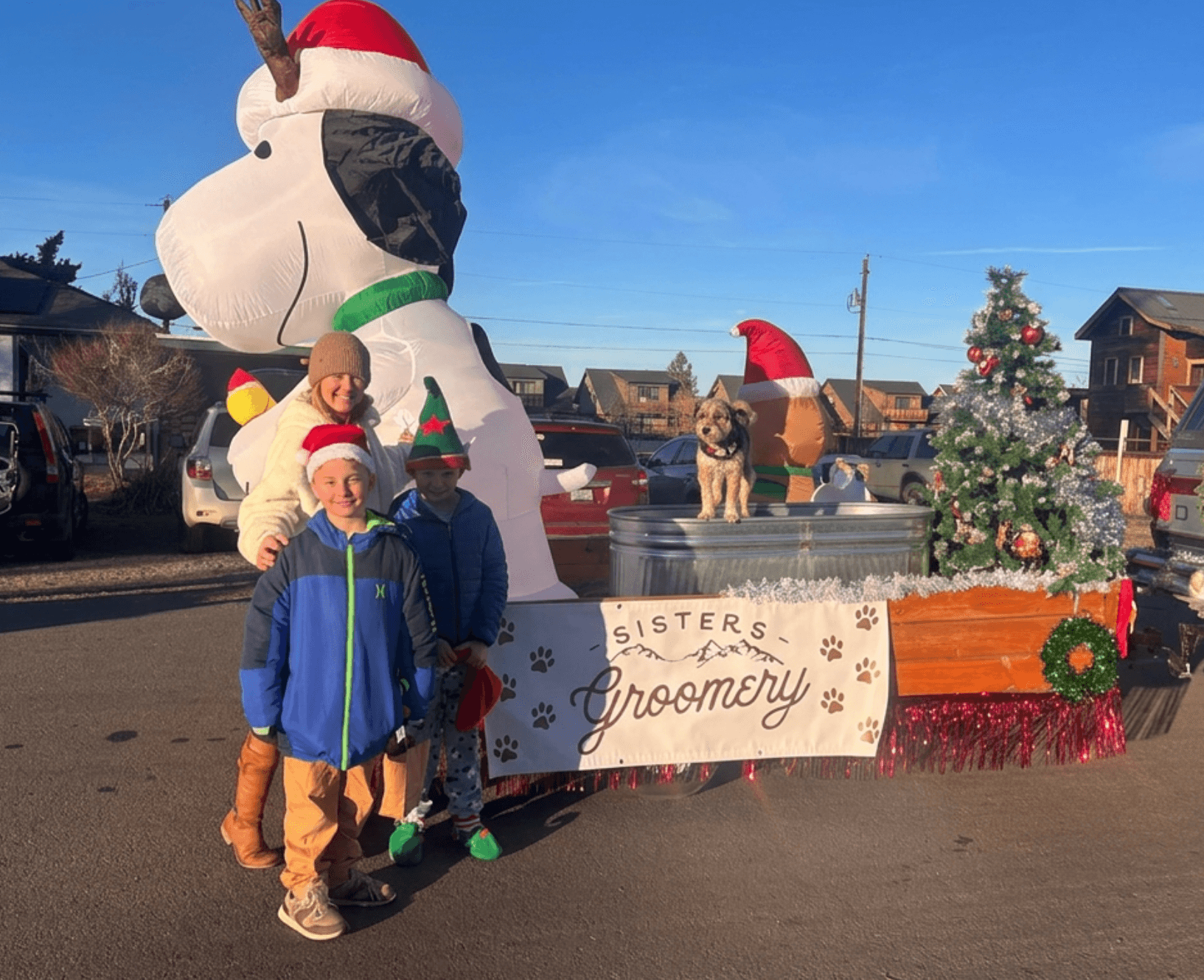 Children in winter clothing and Santa hats stand in front of a Snoopy float with Christmas decorations and a sign that reads 'Sisters Grommly'.