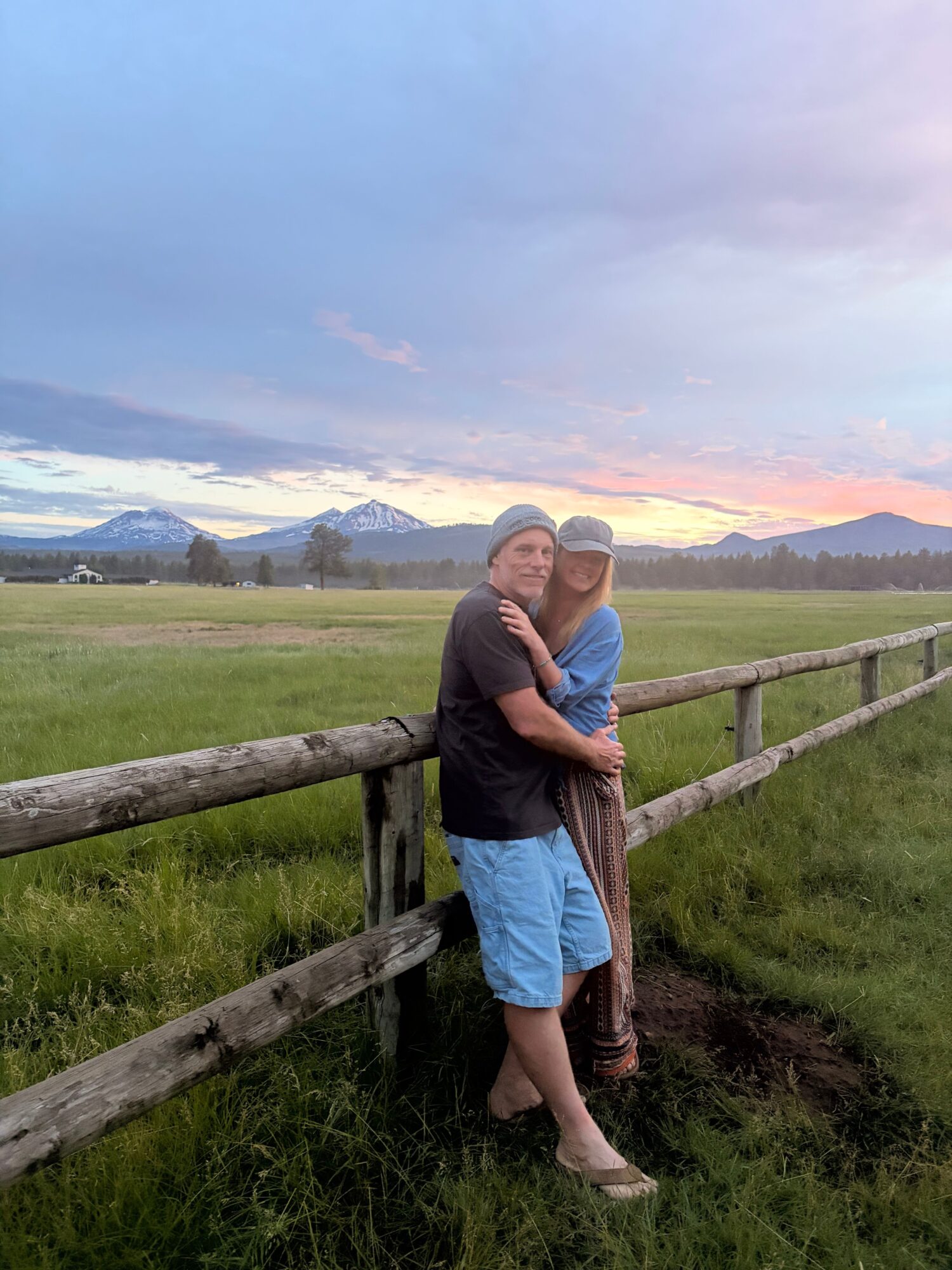 Two people standing on a wooden fence with a landscape and sunset sky in the background.