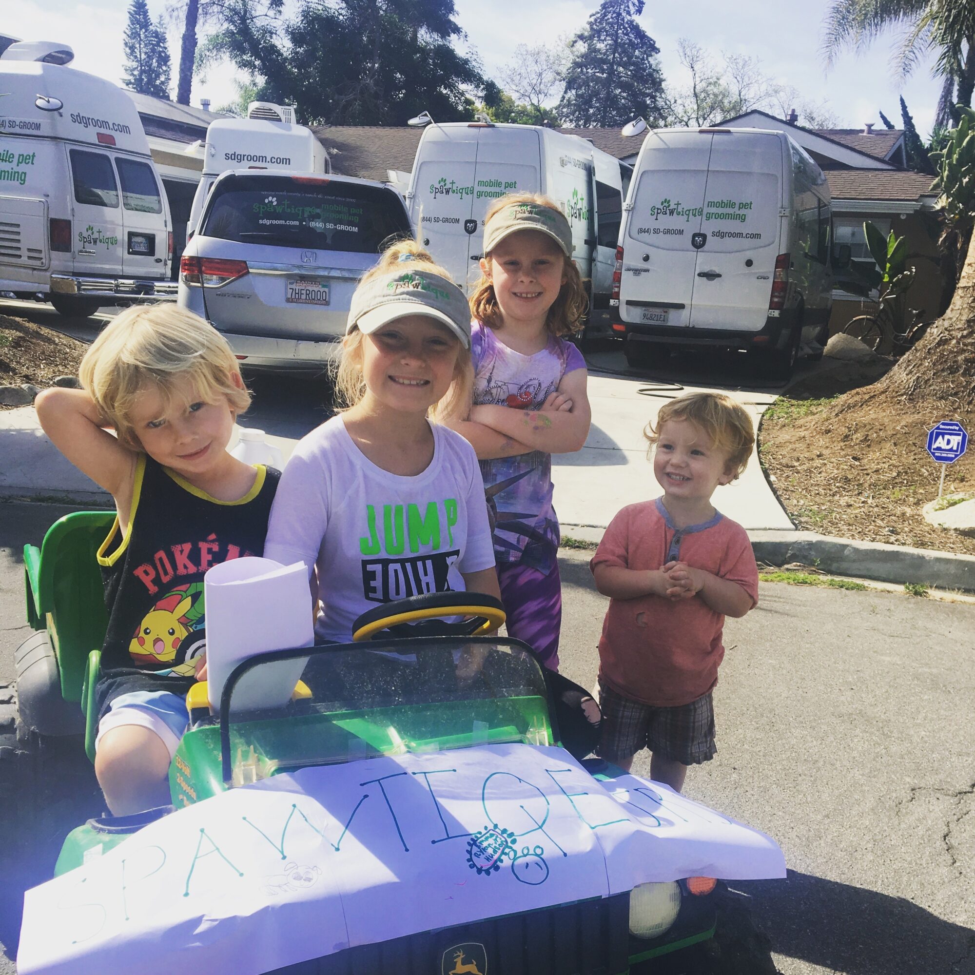 Four children outdoors near parked vehicles, one sitting in a toy car, smiling and posing for the photo.