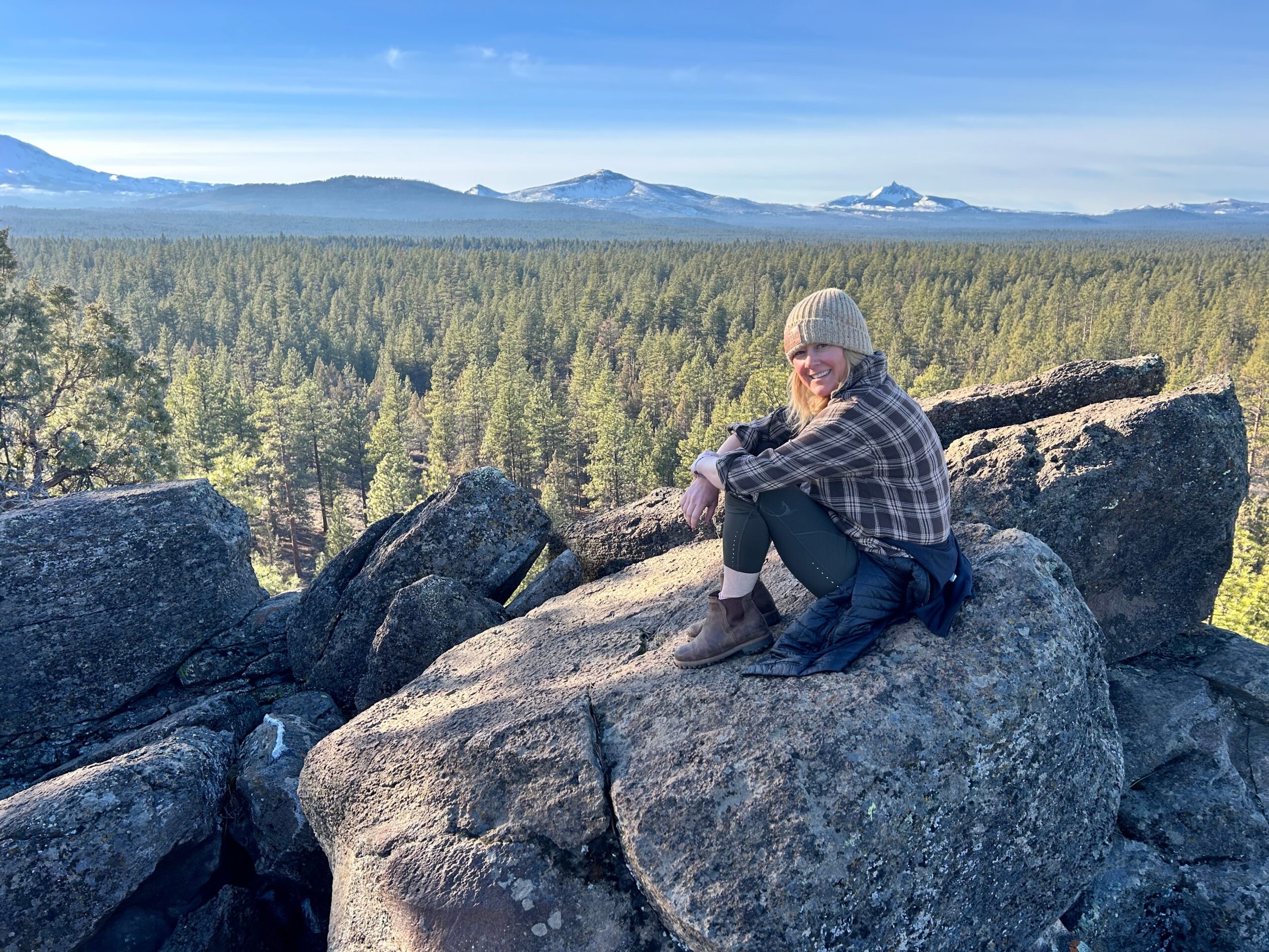 Person sitting on large rocks overlooking a forested landscape with mountains in the distance, wearing a hat and plaid shirt.