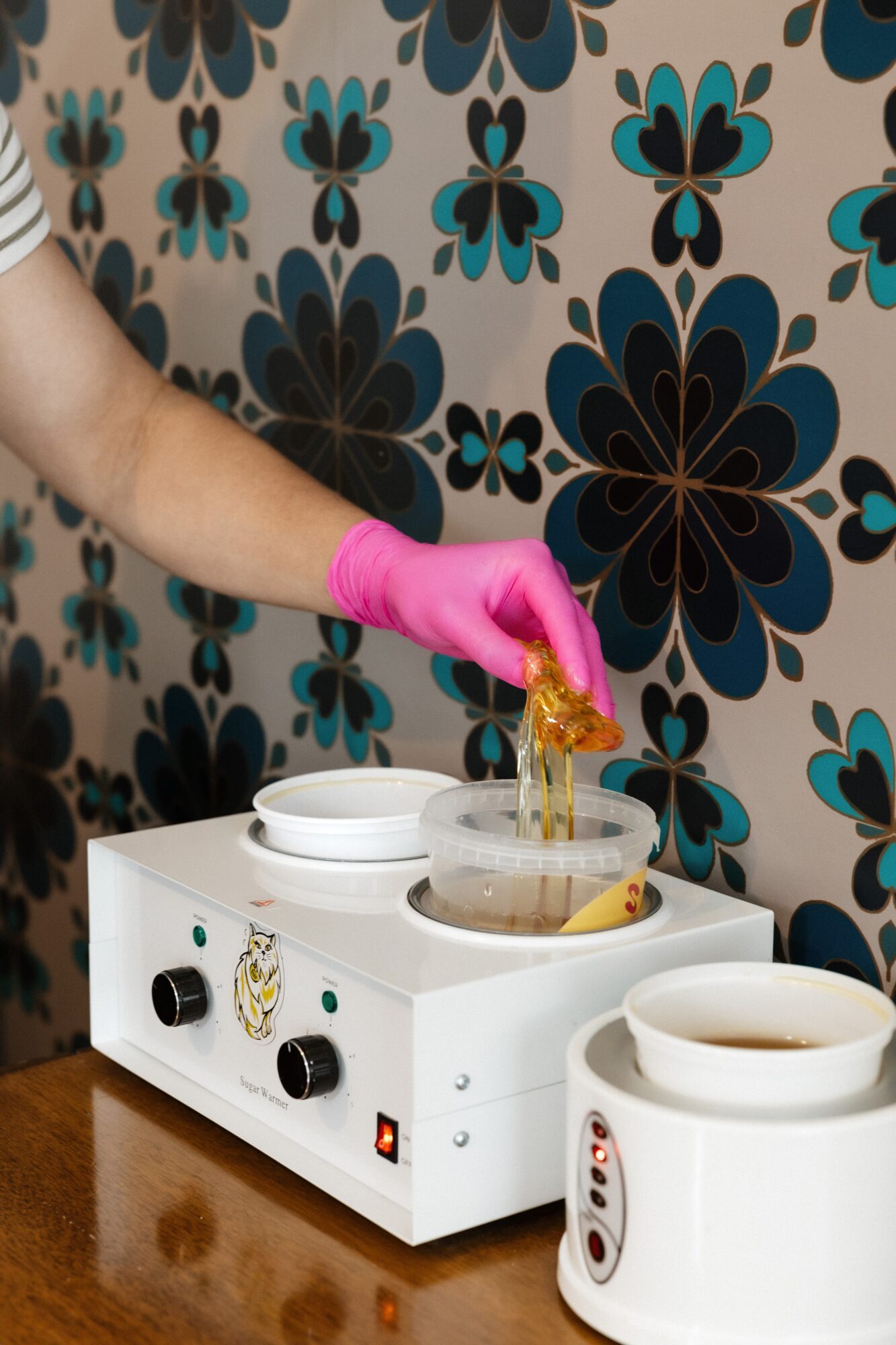 Person wearing pink glove handling a yellow substance over a white heating device with bowls, against a floral patterned wall.