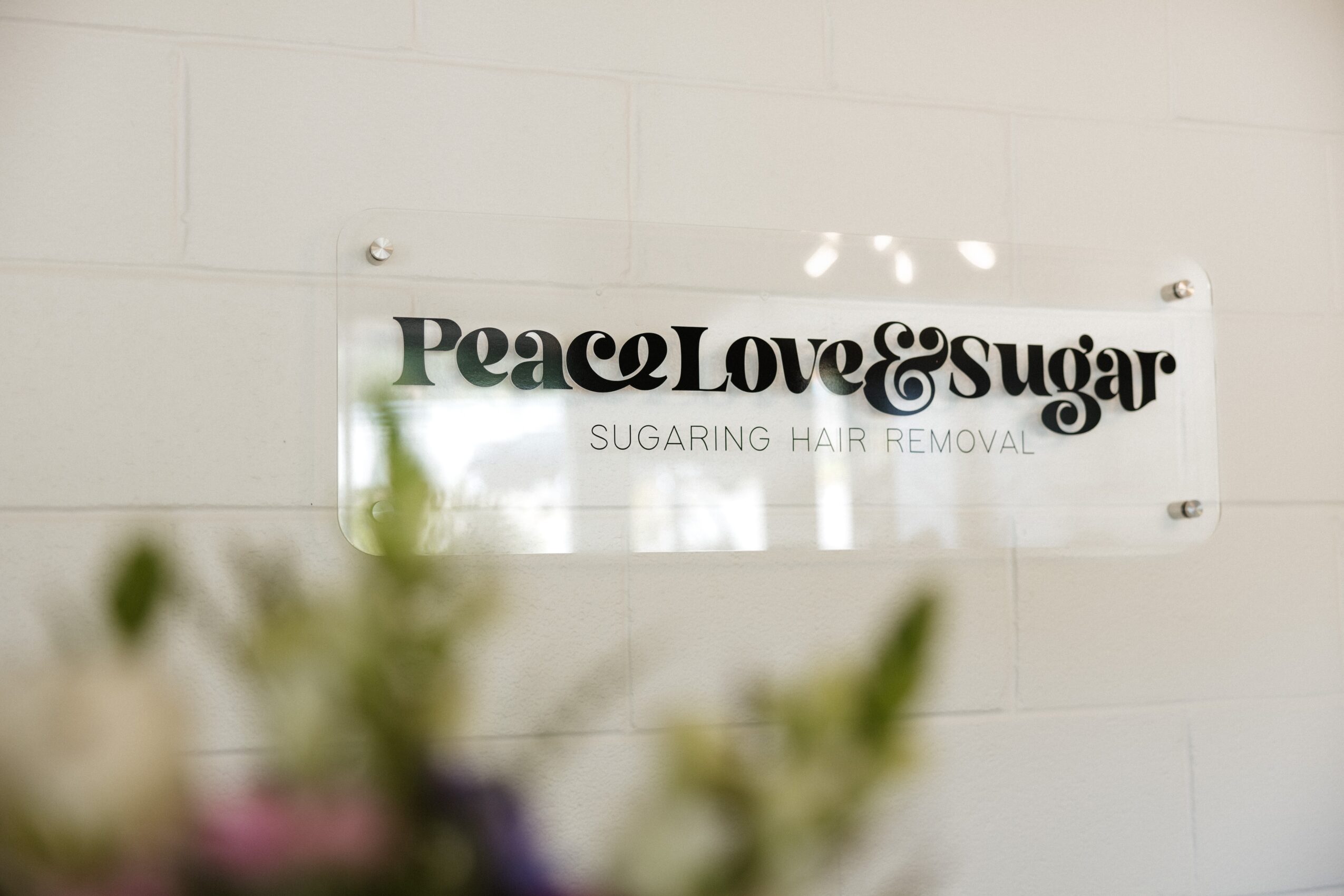 Sign on a wall reads 'Peacock Love & Sugar' with smaller text below, 'SUGARING HAIR REMOVAL', on a white background.