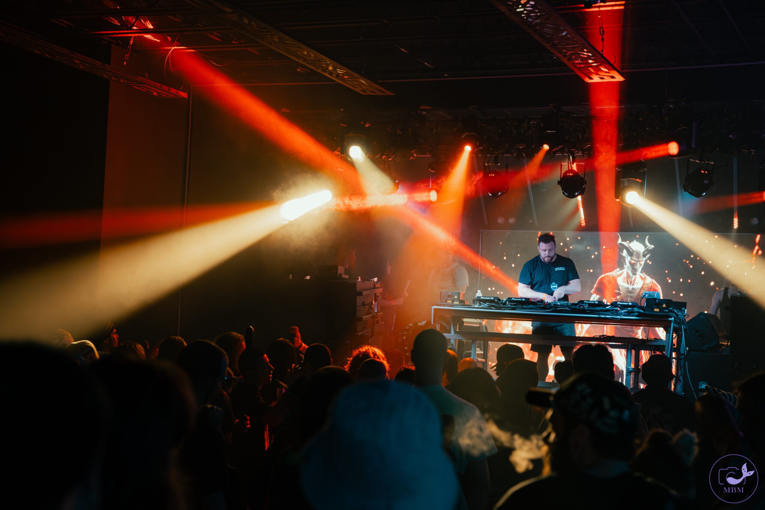 DJs perform on stage with colorful lights shining on an audience in a dark venue.