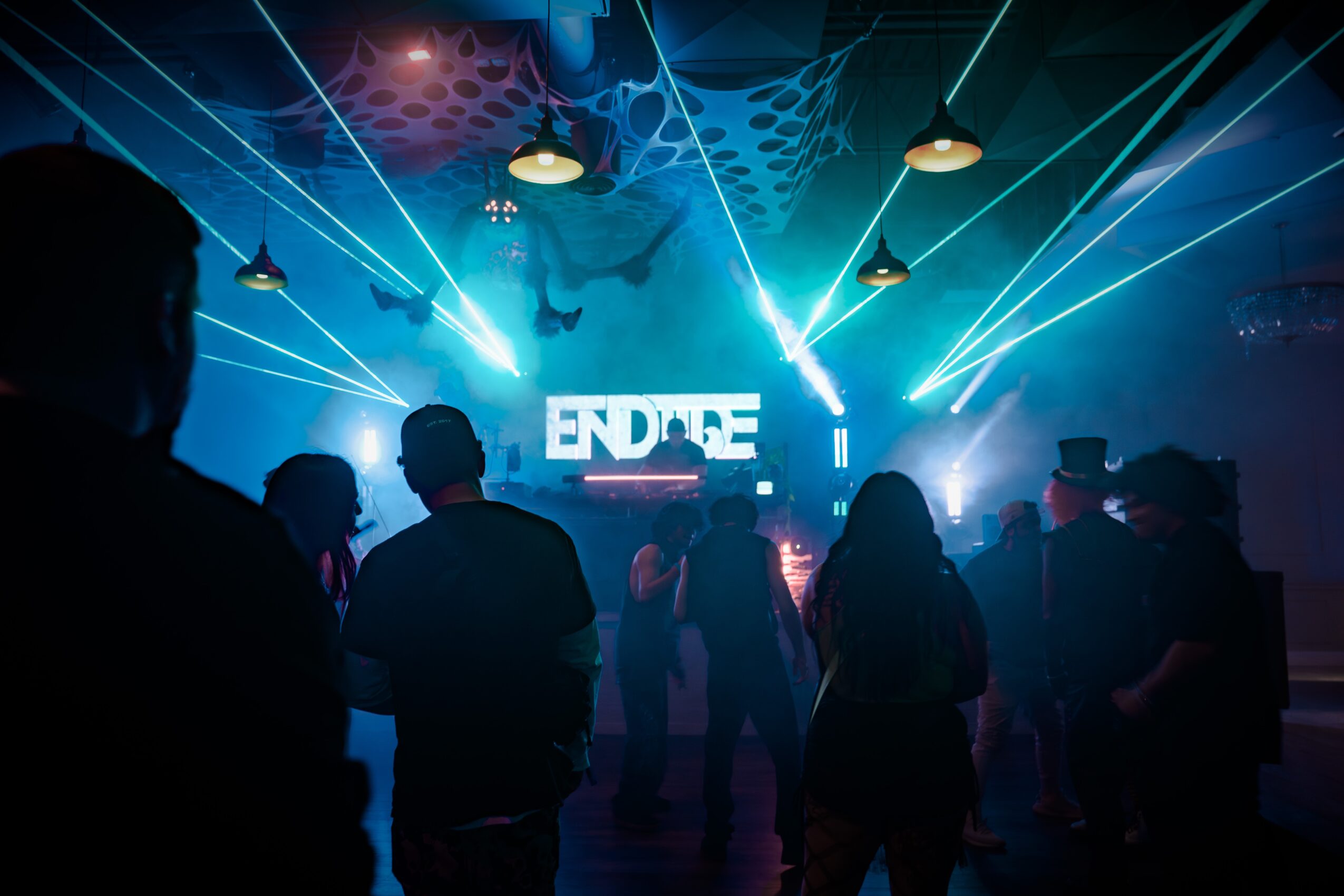 People dancing in a club with blue laser lights and a DJ booth, silhouettes visible against colorful lighting, lively atmosphere.