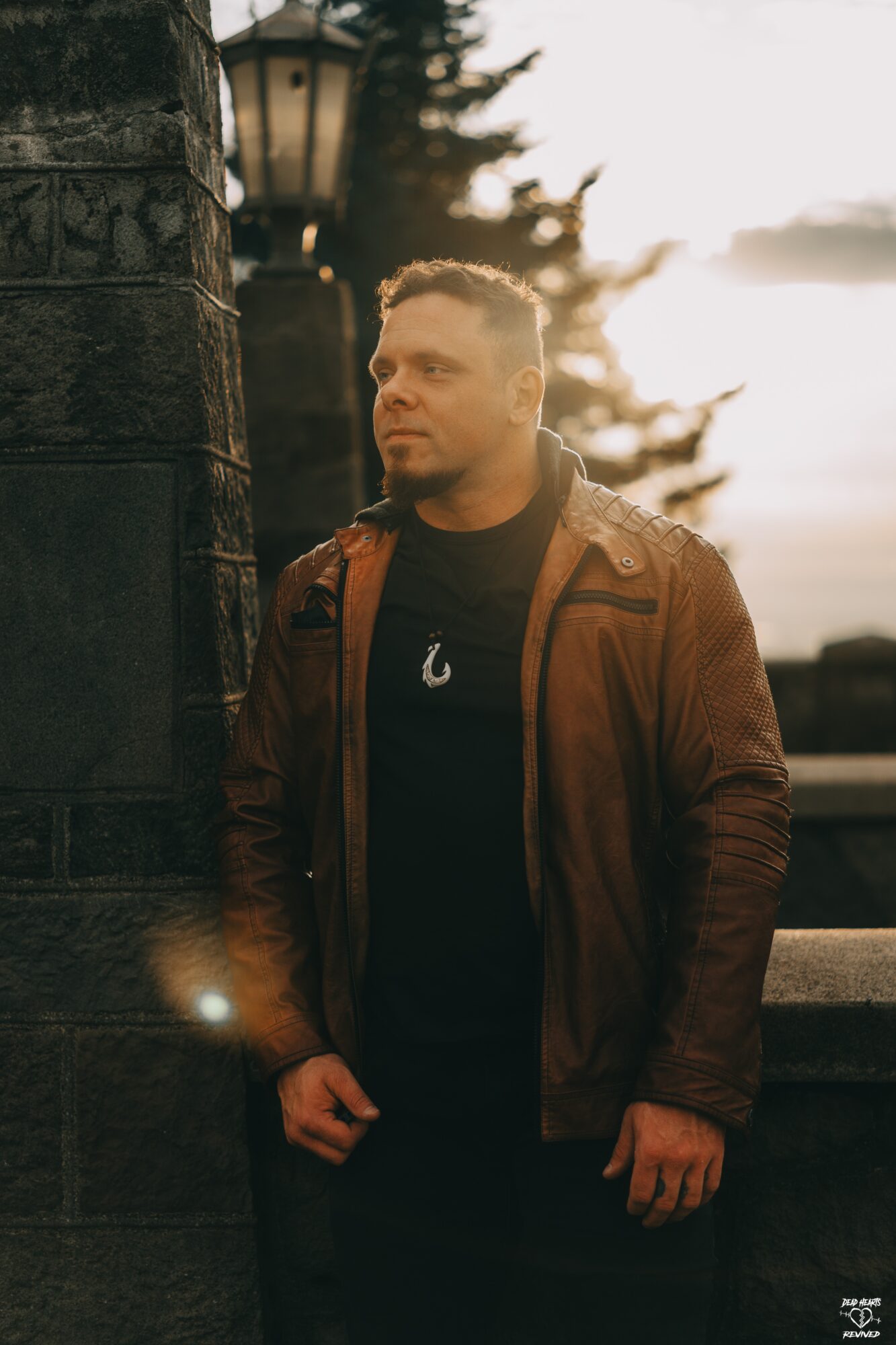 Man standing outdoors near a stone wall with trees and a lamp post in the background, sunset lighting, wearing a brown jacket.