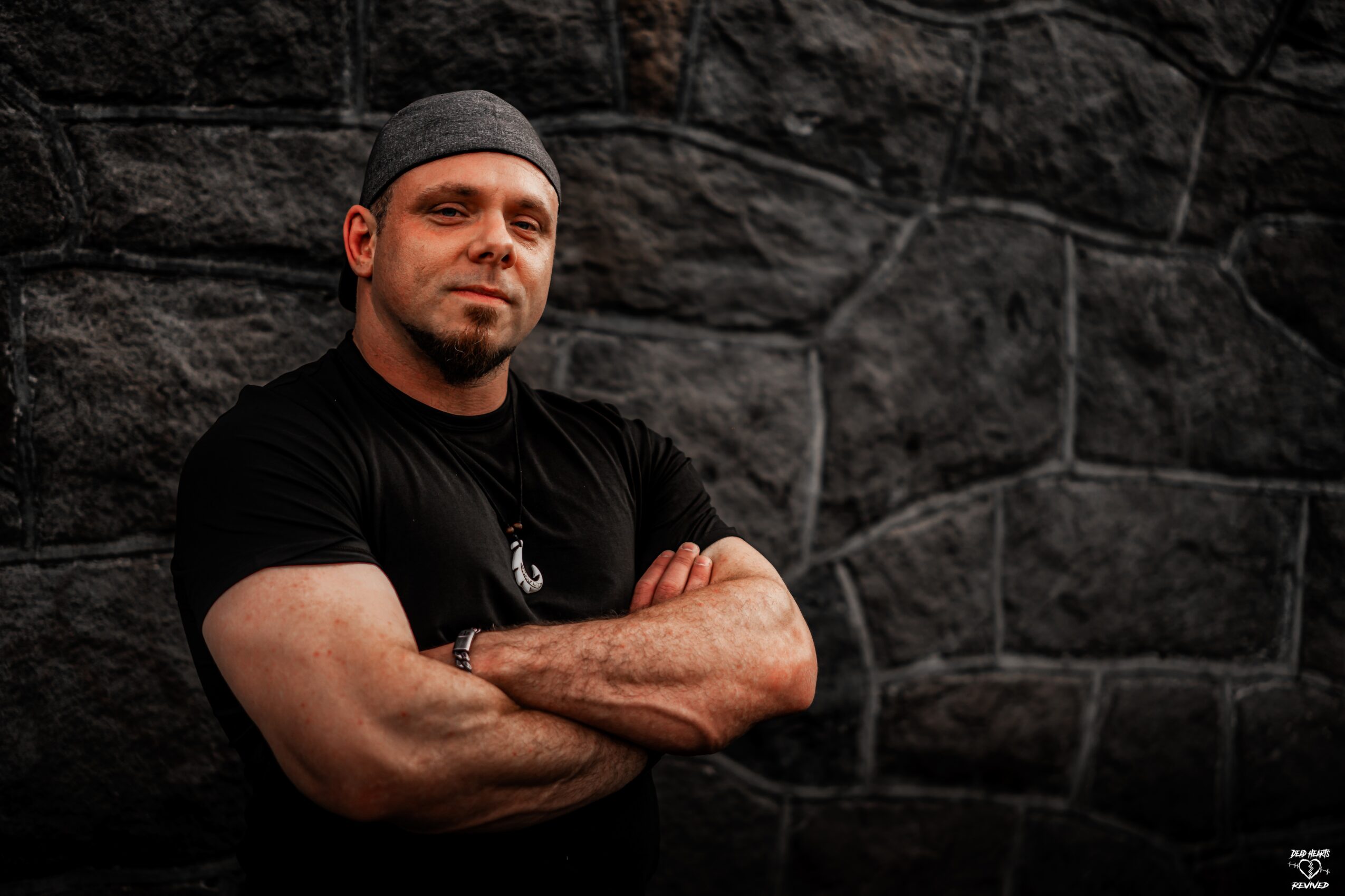 Man with arms crossed, wearing a black shirt and headband, standing against a stone wall.