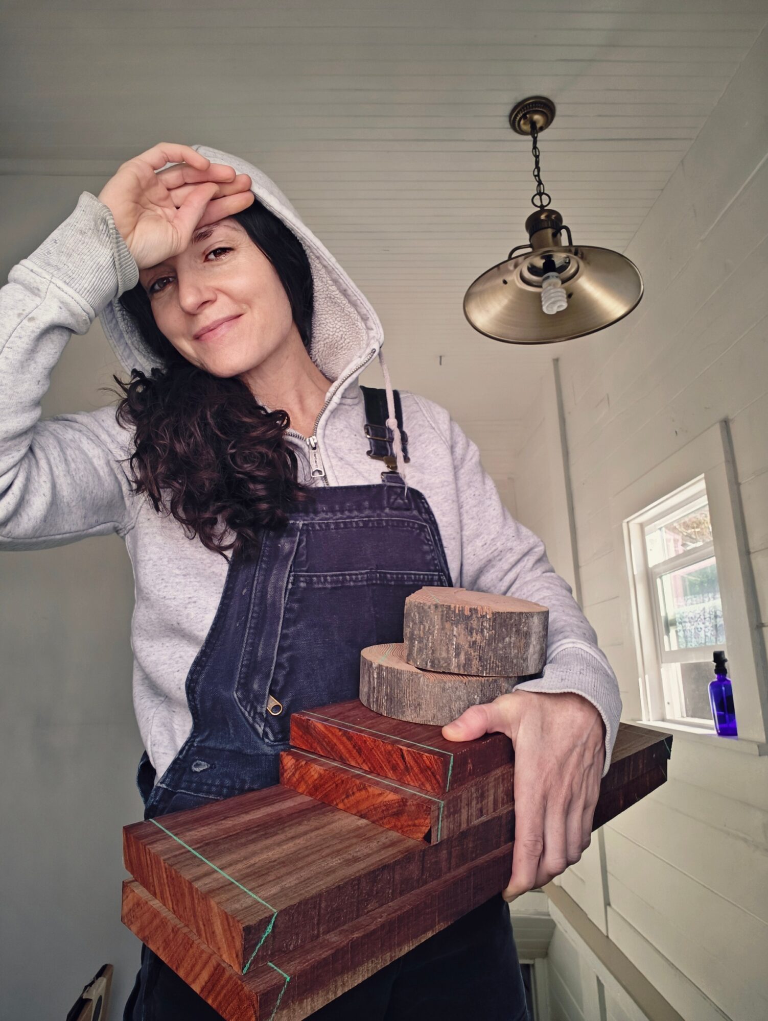 Woman with dark hair in a hoodie and apron holding wooden blocks indoors, with a window and ceiling light visible.