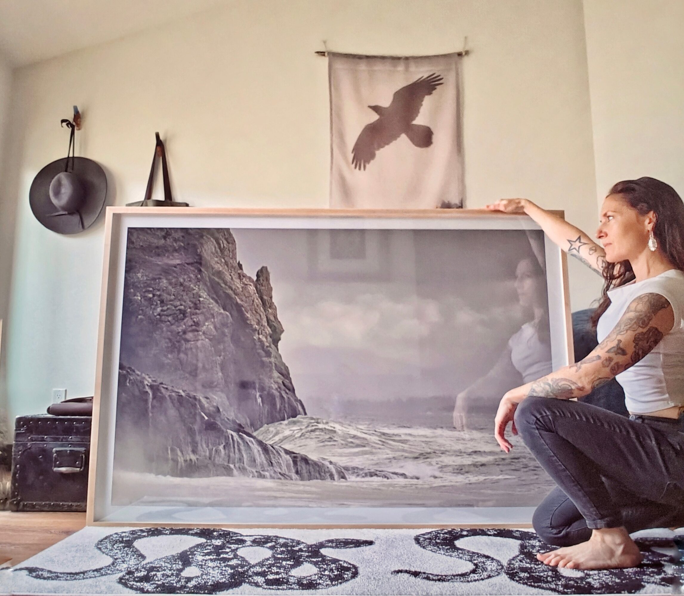 Person with tattoos sitting on floor holding large framed landscape photograph of rocky coast and cliffs.