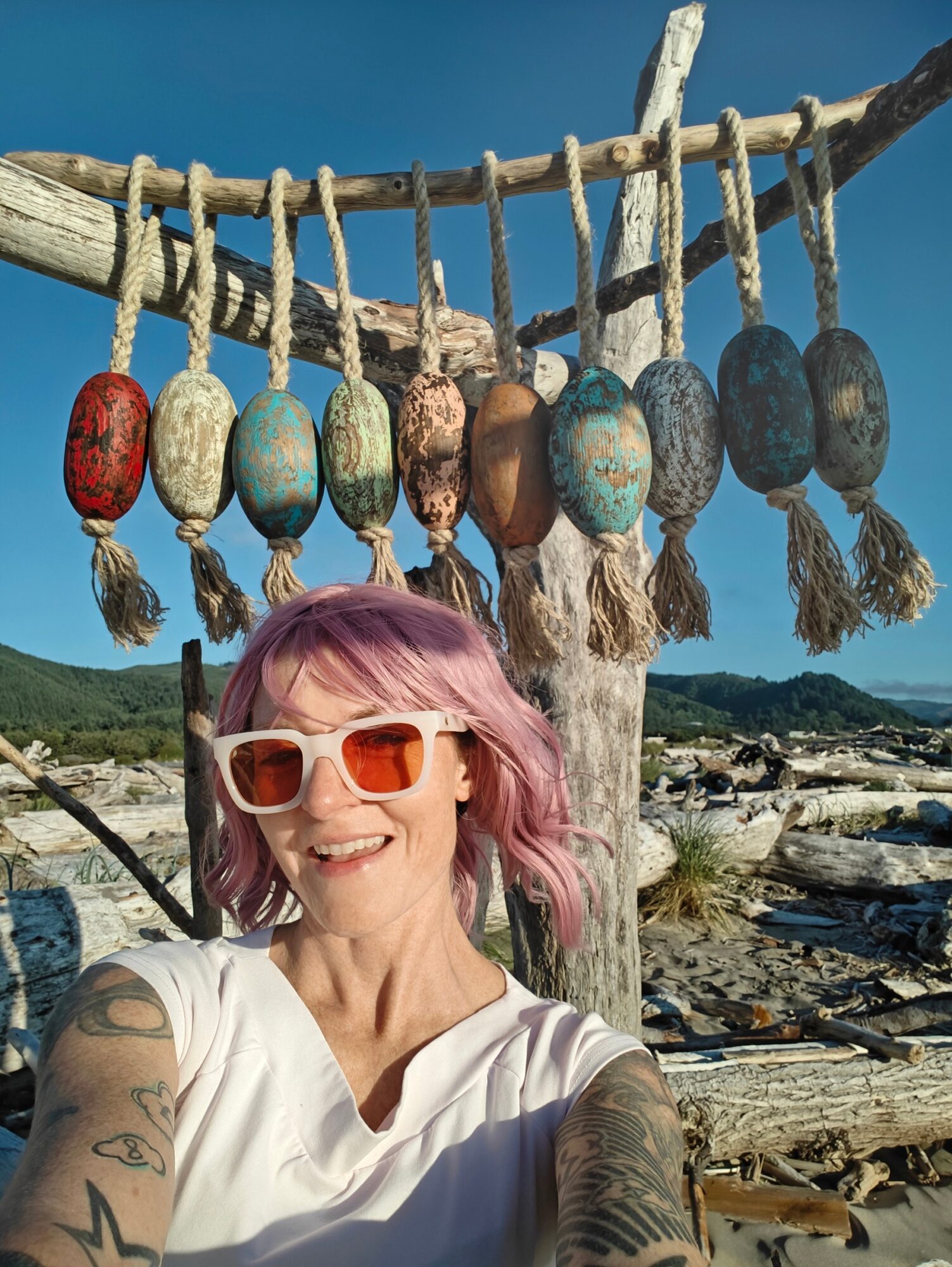 Woman with pink hair and sunglasses smiling outdoors near a tree with hanging painted rocks, landscape background.