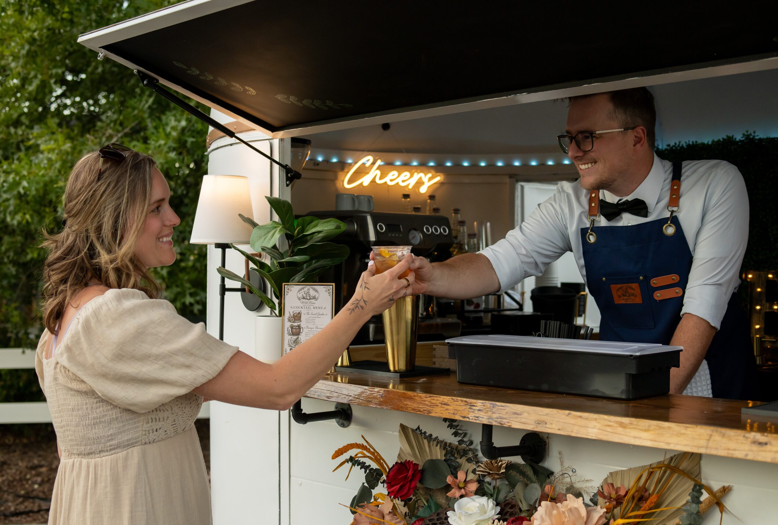 A woman receives a drink from a man at a bar with a neon sign reading 'Cheery' and floral decorations.
