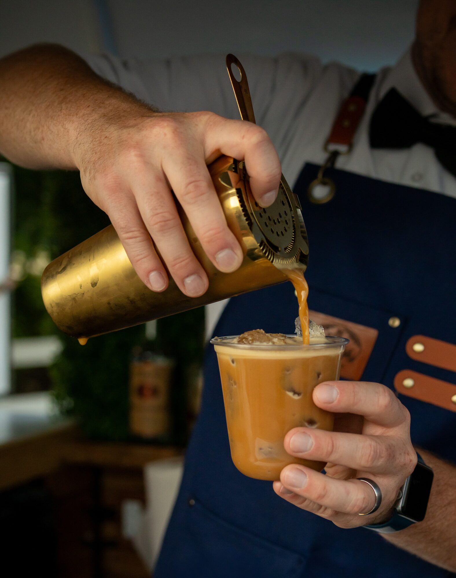 Barista pours coffee from a gold-colored container into a clear cup, wearing a uniform with a bow tie and apron.