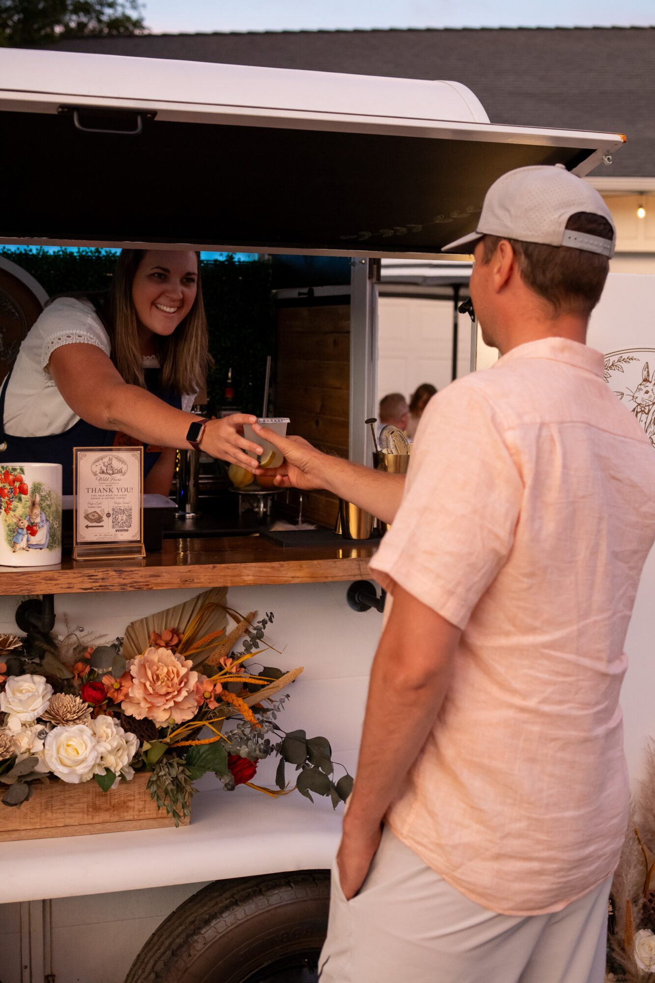 A person in a light-colored shirt and cap receives a drink from a smiling woman behind a counter at an outdoor event.