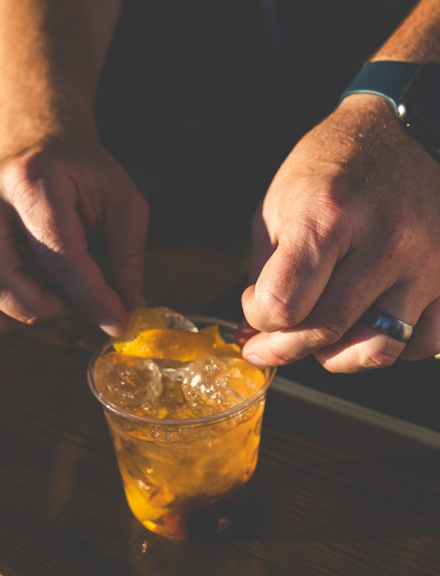 Hands preparing a drink with ice and orange slices in a glass, on a dark surface.