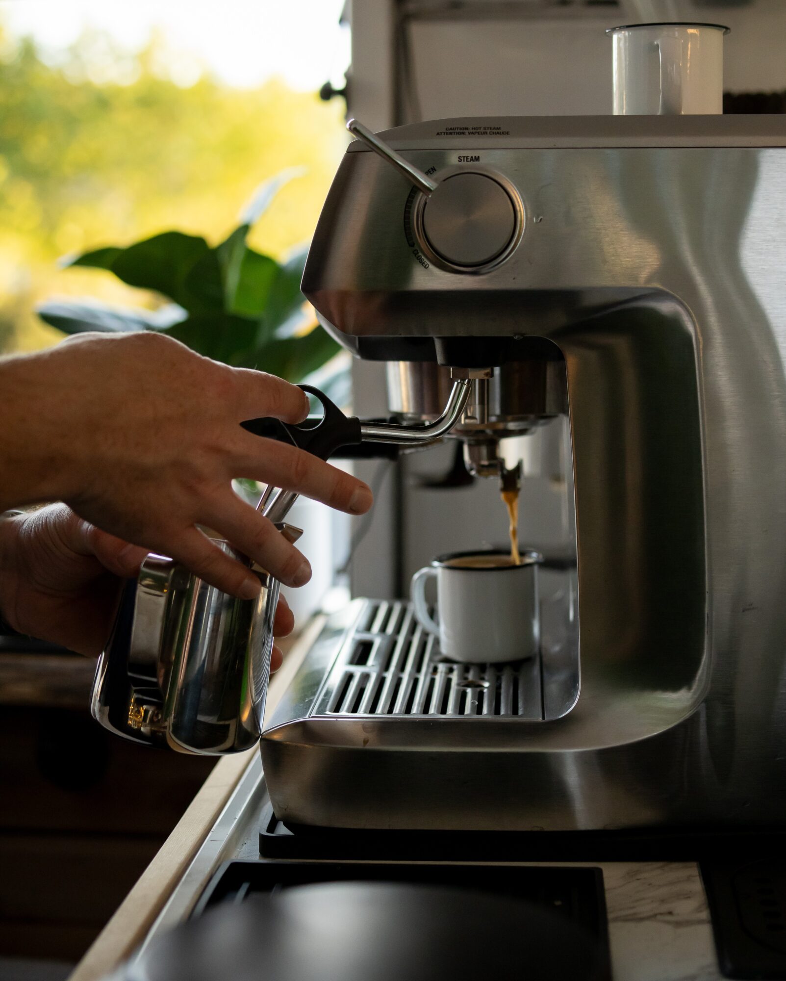 Person using an espresso machine to brew coffee into a small cup, with a hand holding a milk frothing pitcher, in a kitchen setting.