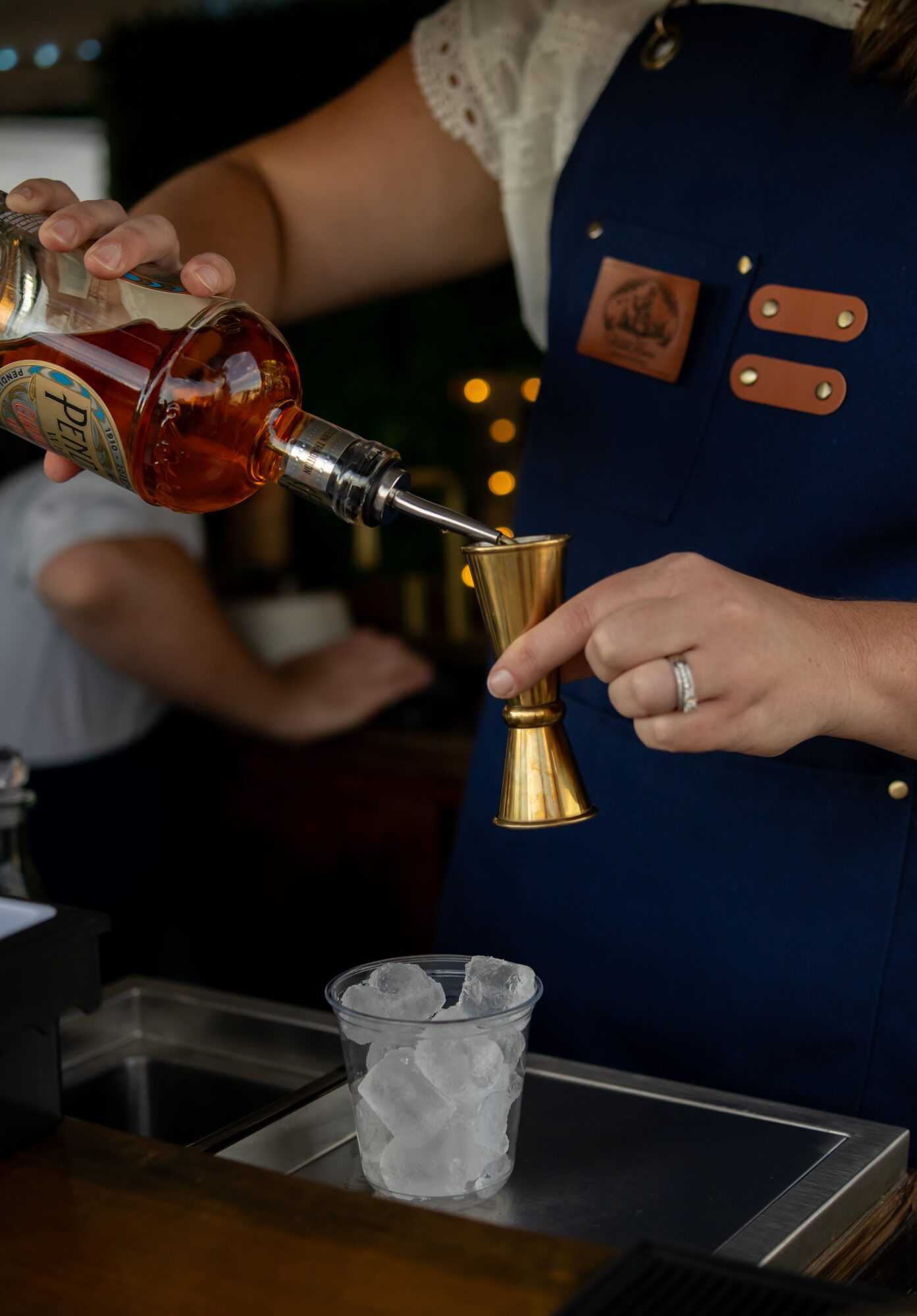 Person pours a drink from a bottle into a jigger over a glass with ice, wearing a blue apron. 20 words.
