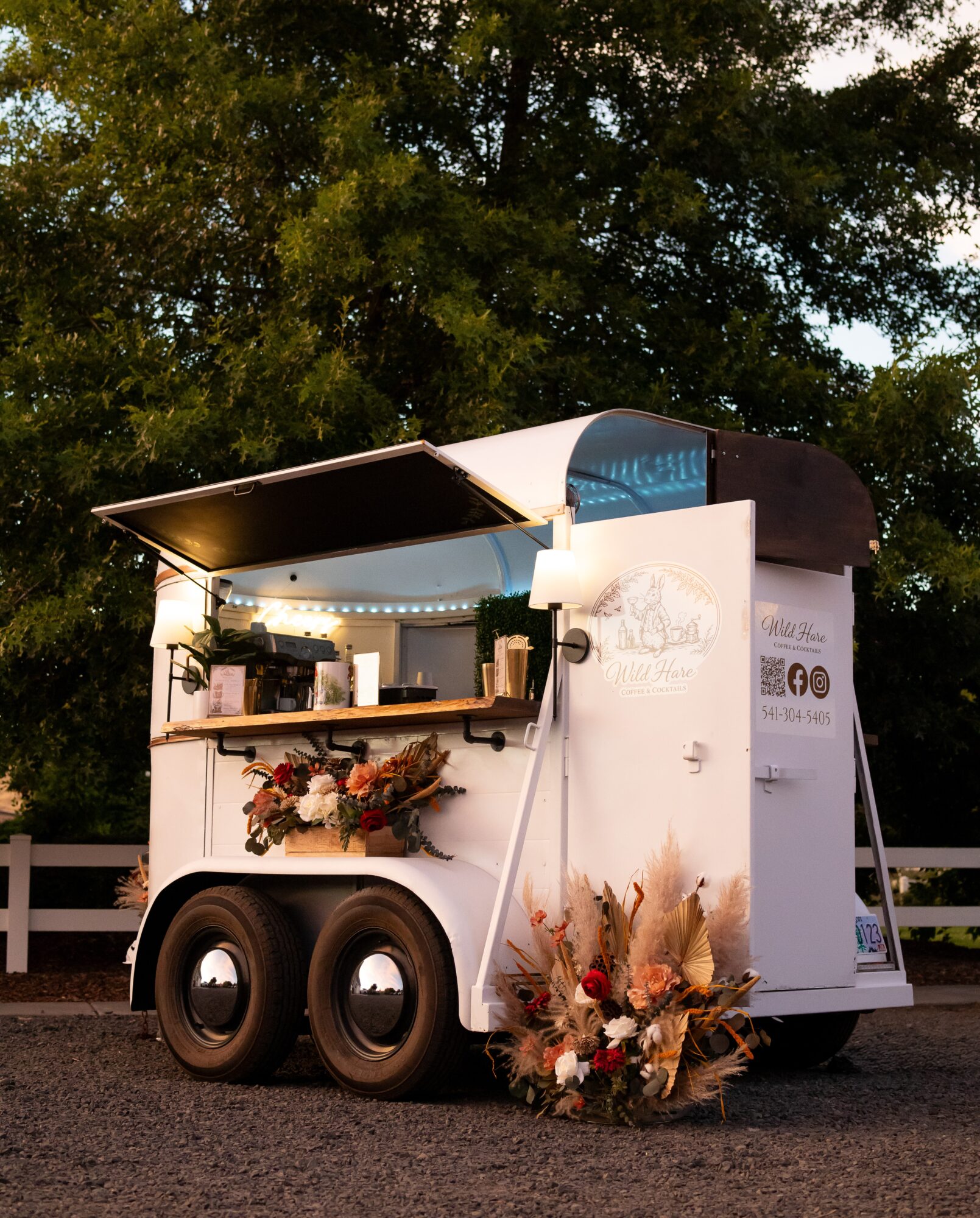 Food truck with open window, decorated with flowers, parked outdoors with trees in background.