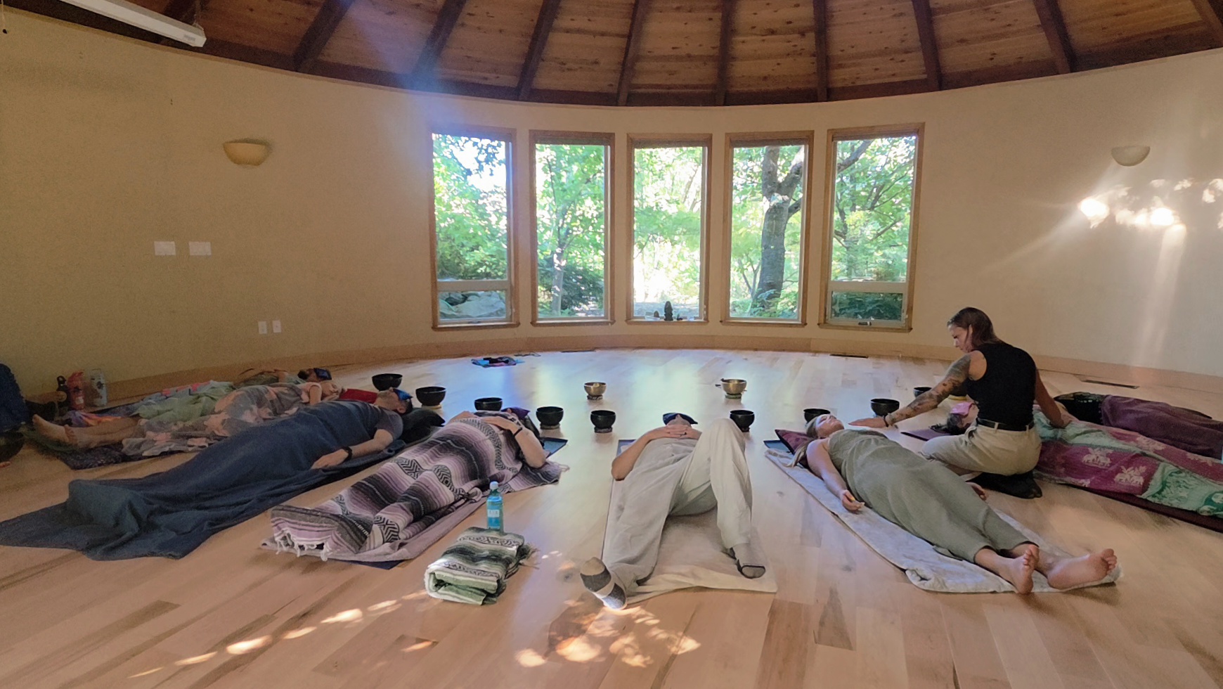 People lying on mats in a circular room with large windows and wooden ceiling, some resting, some sitting, surrounded by personal items.