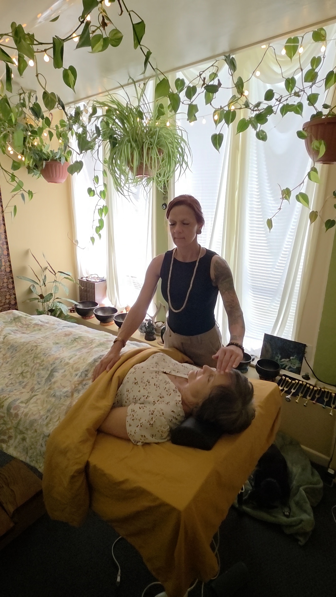 A woman receiving a massage in a room with hanging plants and natural light, lying on a yellow-covered table.