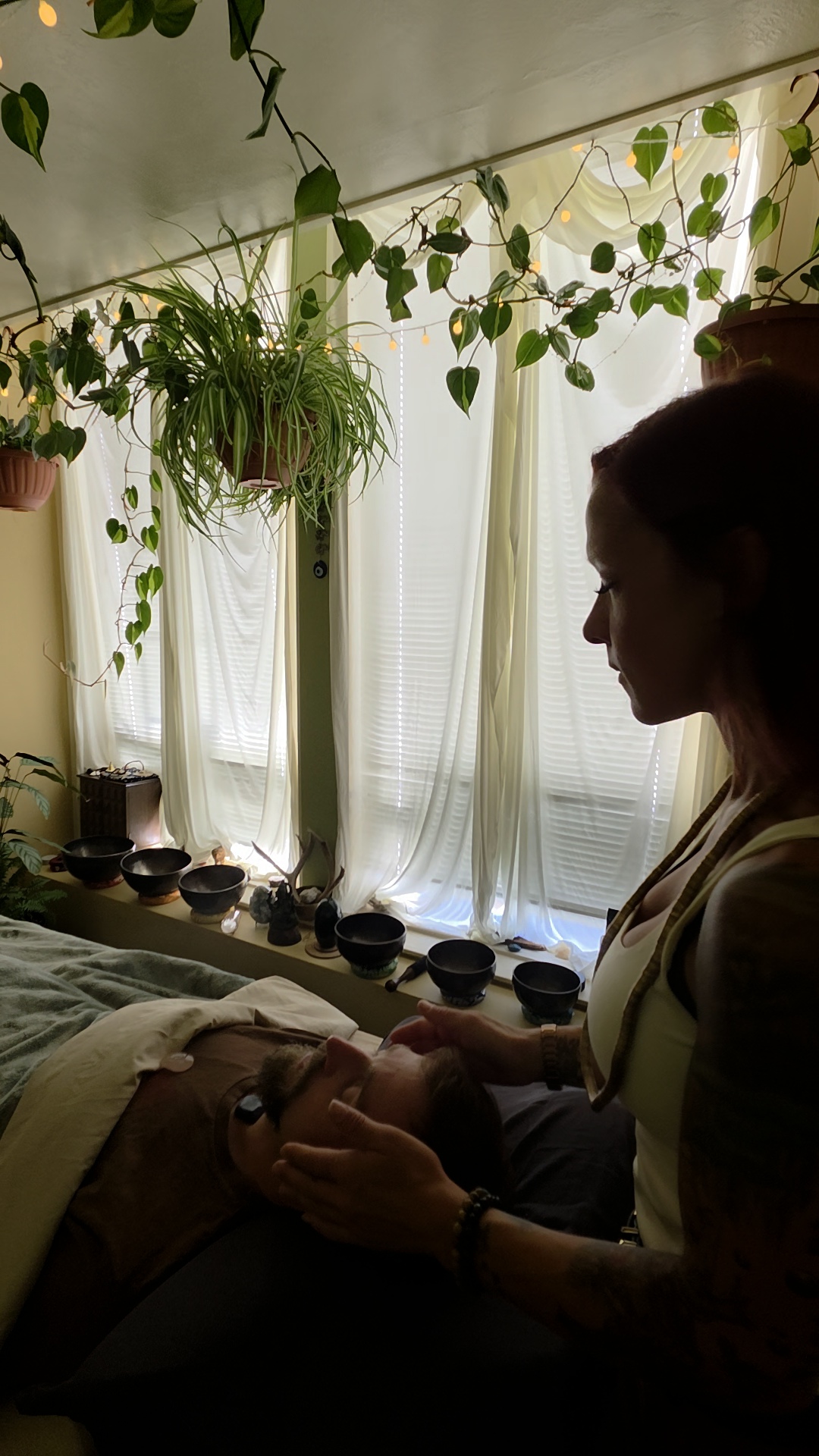 Person standing near a window with hanging plants and sheer curtains, holding a small object, in a dimly lit room.