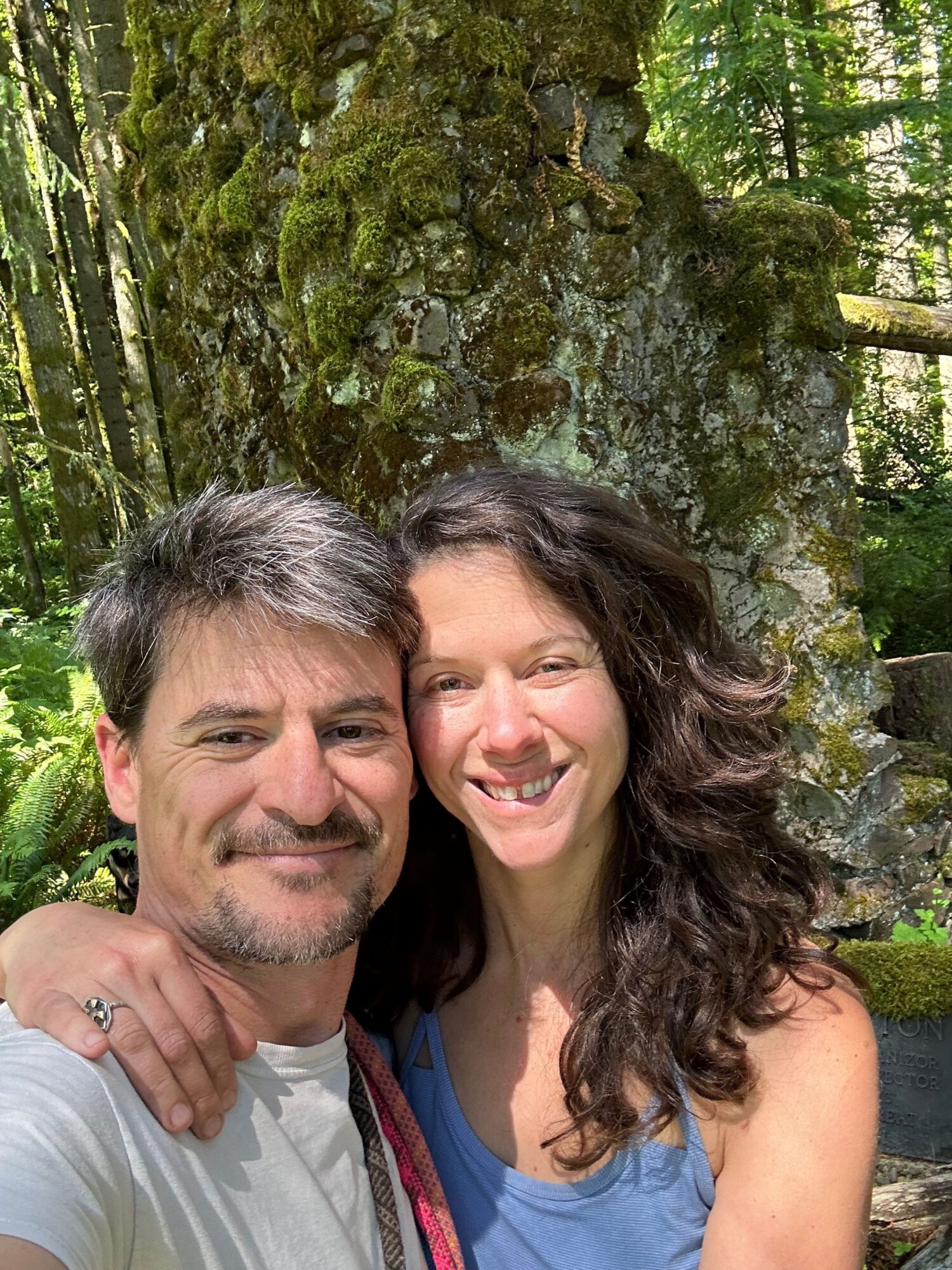 Two people smiling outdoors near a moss-covered tree, one with curly hair and the other with straight hair, both looking at the camera.
