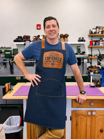Man wearing a blue apron with orange straps, standing in a workshop with tools and shoes on shelves behind him.