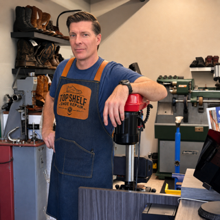 Man in a workshop wearing a blue apron, holding a red and black power drill, surrounded by tools and equipment.