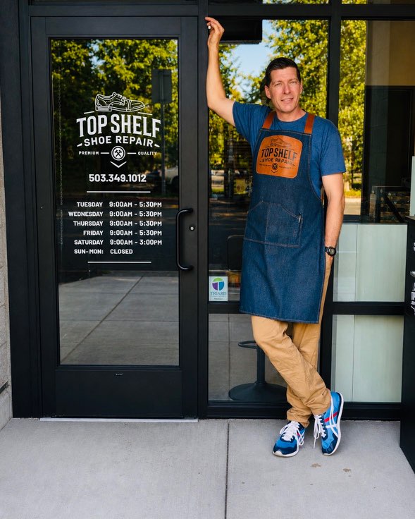 Man in apron leaning against glass door with store sign, standing outside store entrance, smiling, with trees in background.