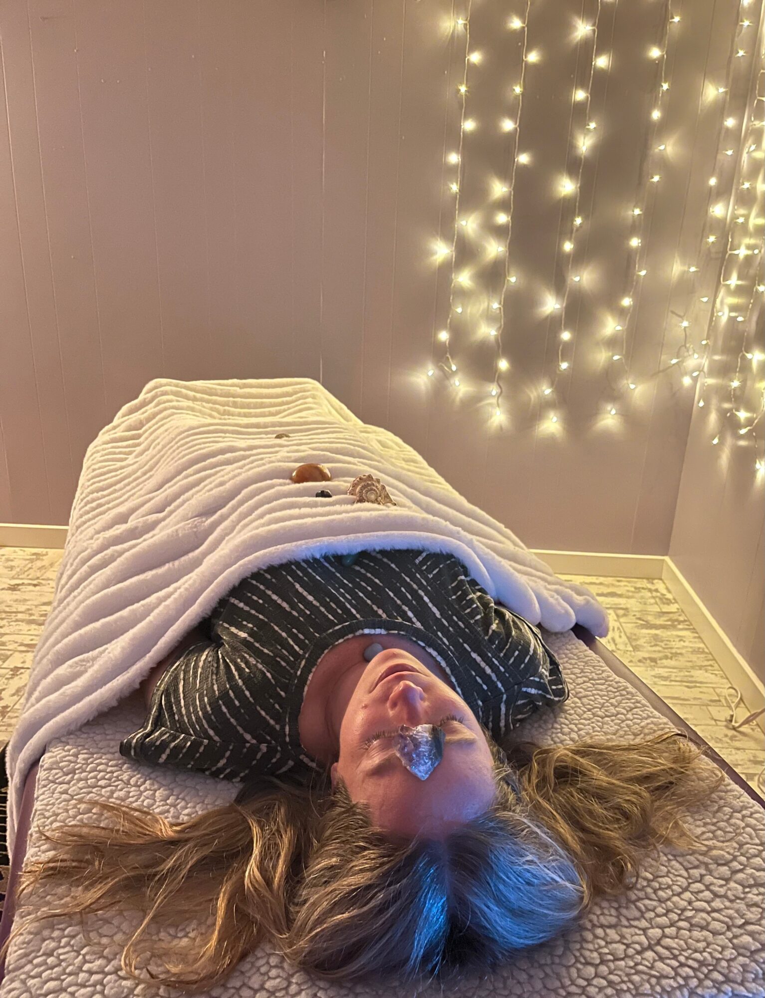 Person lying on a bed with a blanket, head tilted back, in a room with string lights on the wall.