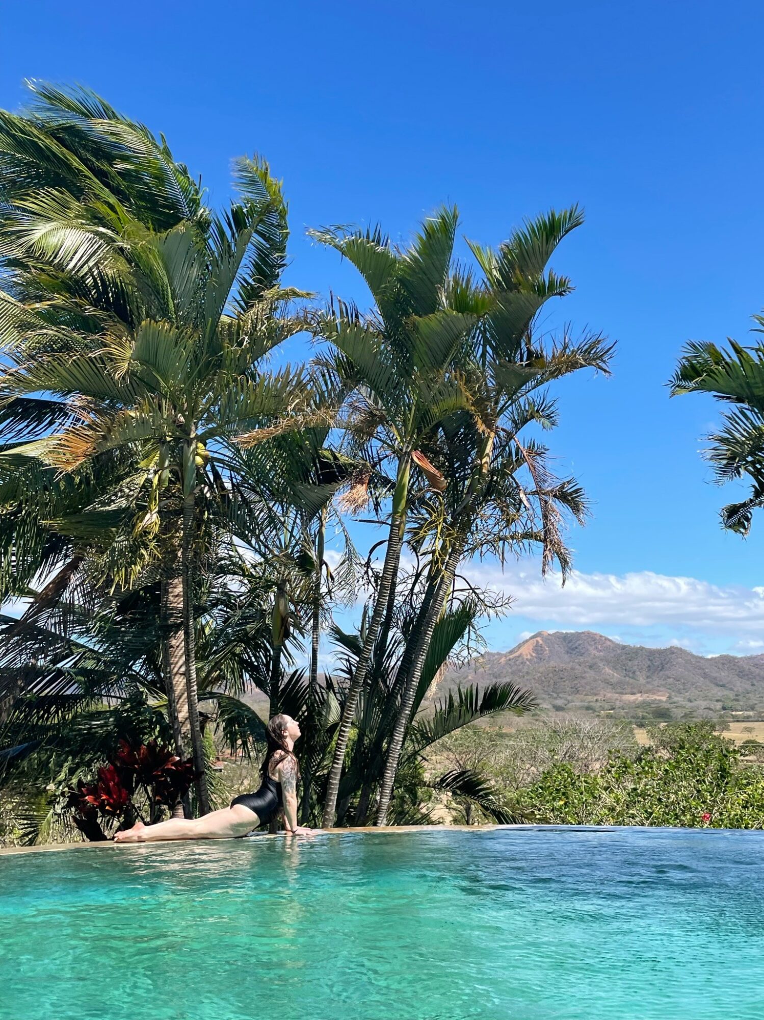 Person sitting on the edge of a pool with palm trees and mountains in the background under a clear blue sky.