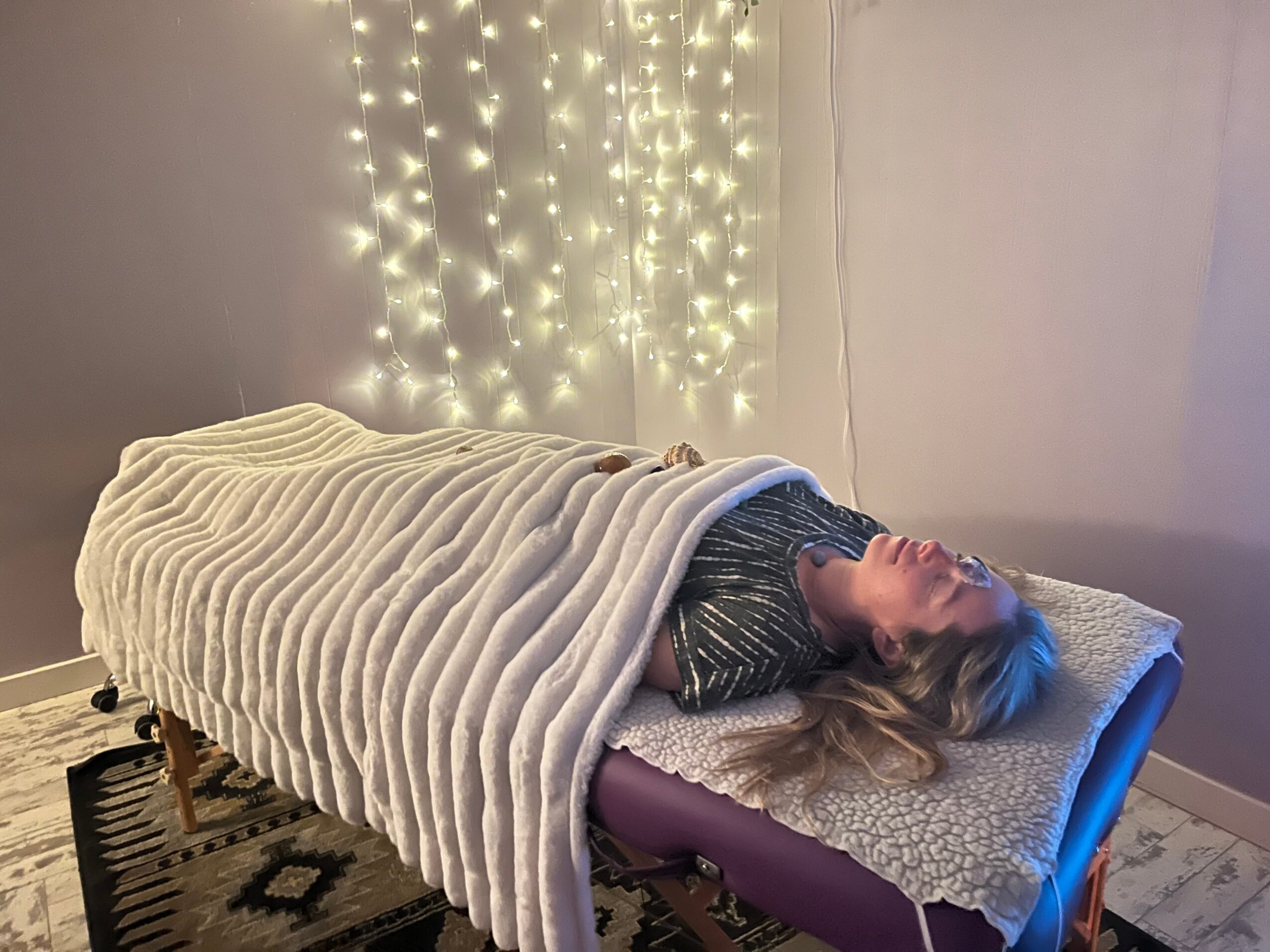 Person lying on a bed covered with a blanket, head resting on a pillow, with string lights on the wall behind.