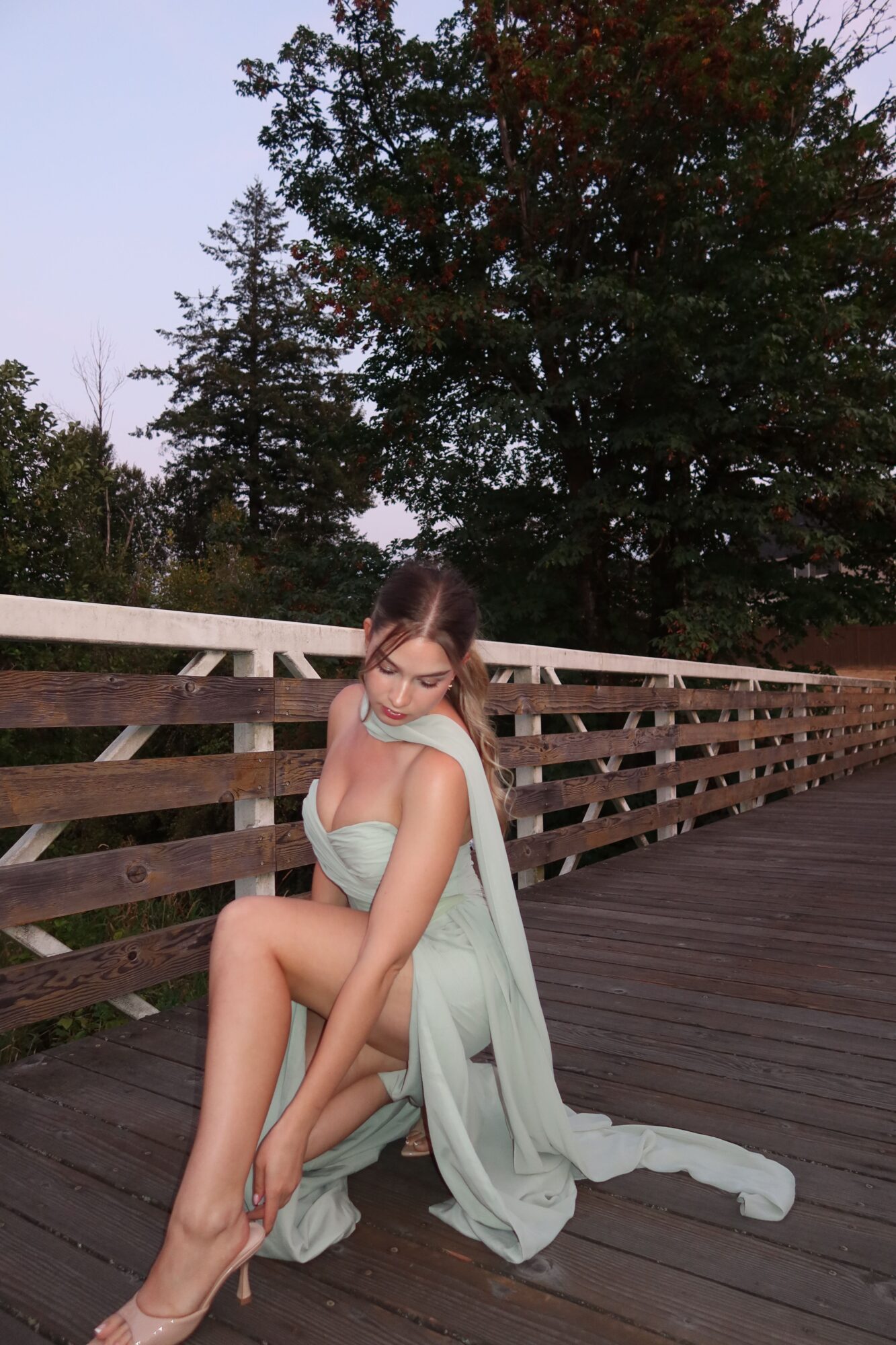 Woman in a flowing dress sitting on a wooden deck, leaning against a railing, with trees in the background.