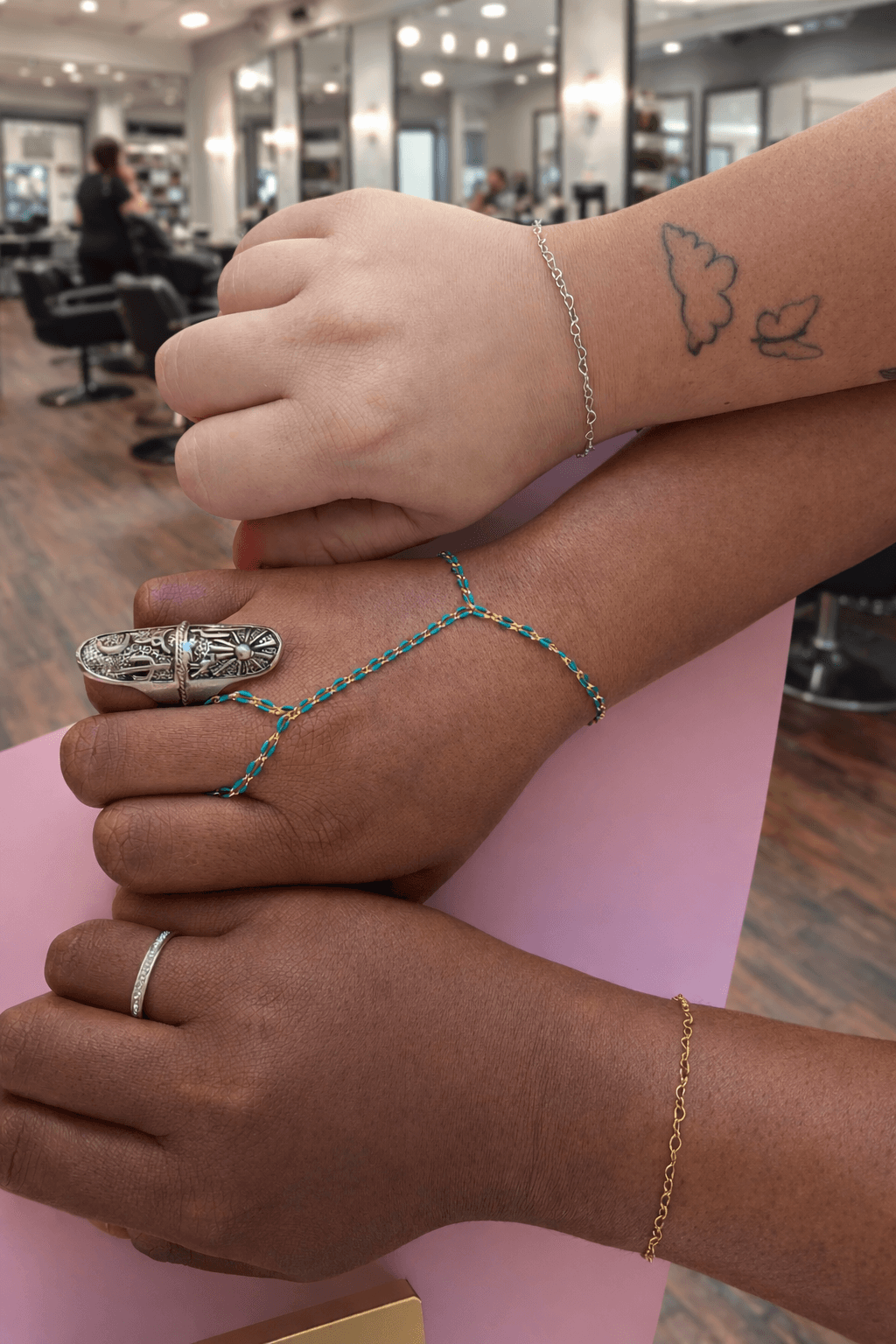Two hands with jewelry and tattoos, one resting on the other, in a salon or similar setting.