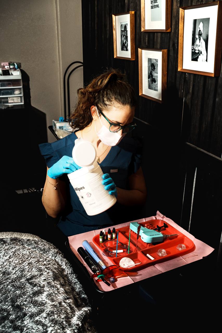 Person wearing glasses and gloves holding a container, working with small bottles on a red tray, in a room with framed photos.