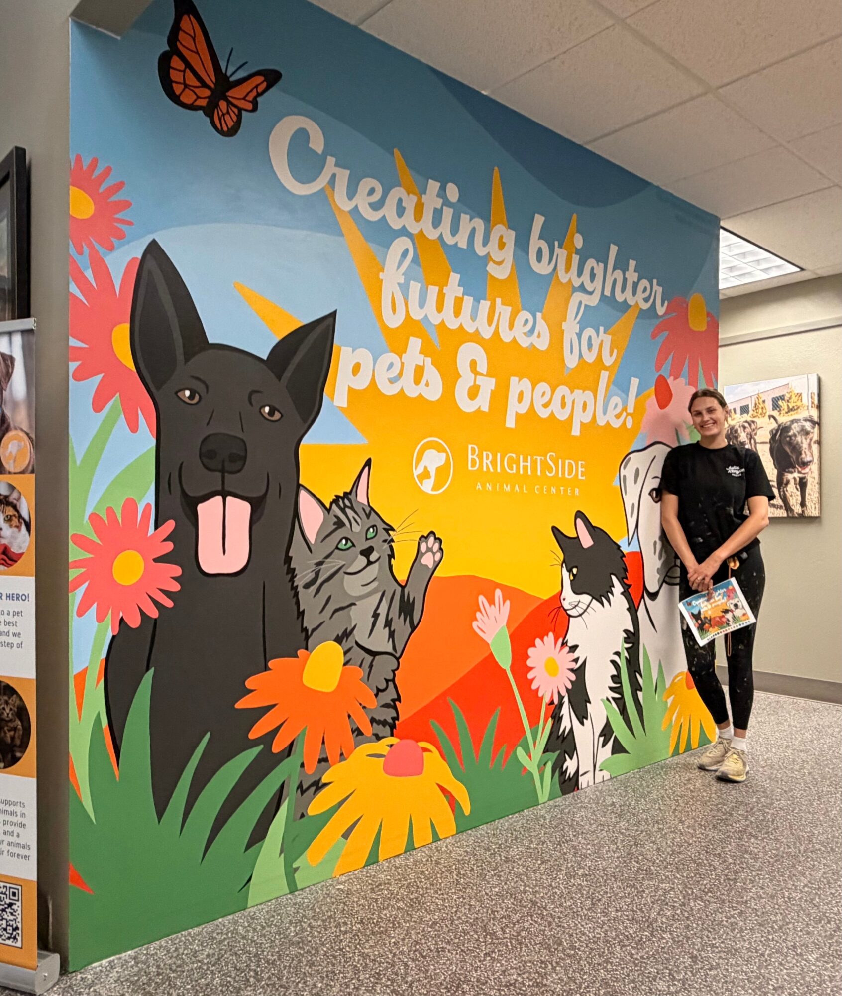 Person standing next to a colorful wall mural with a dog, cat, flowers, and text about creating a brighter future for pets and people.